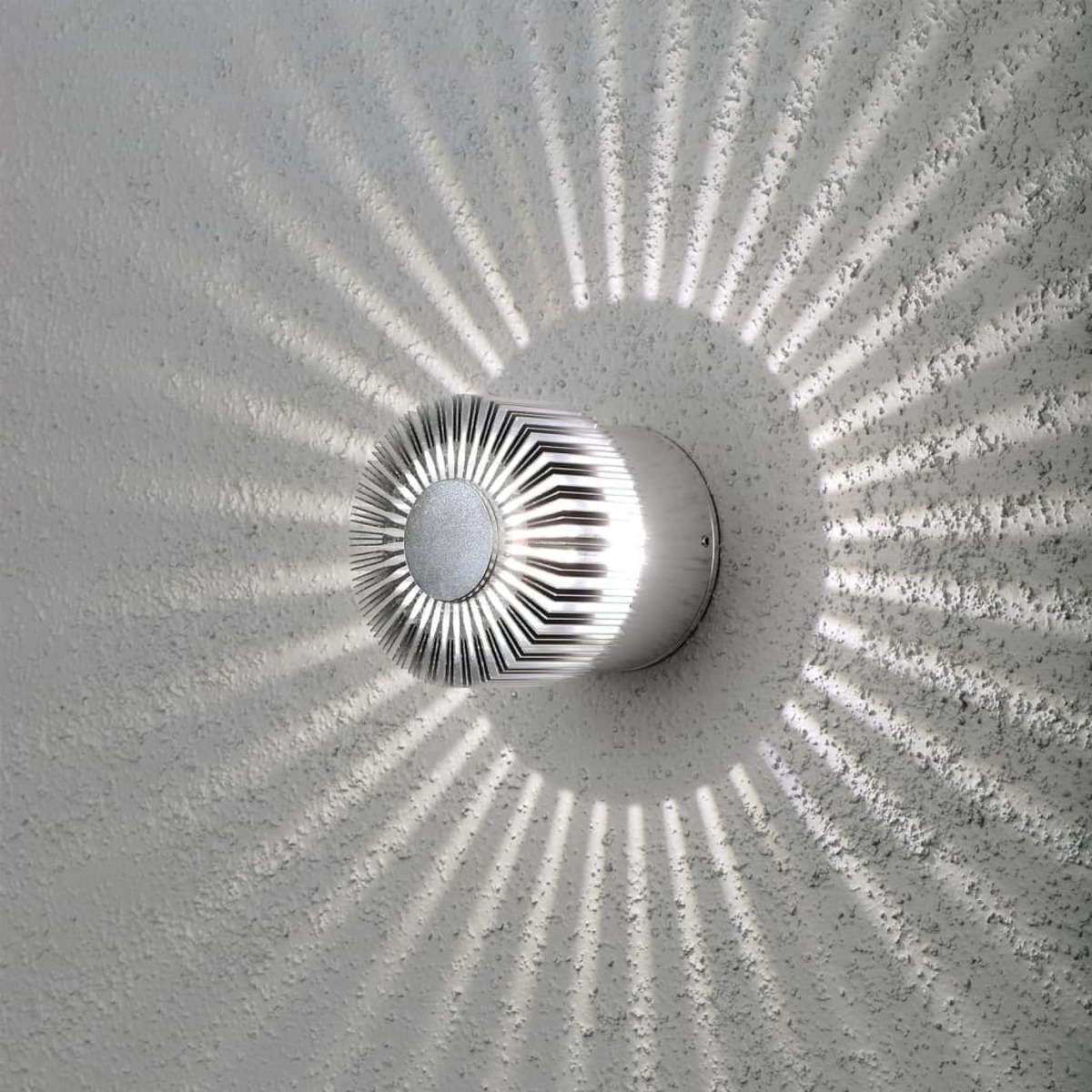 Lampada da parete rotonda argentata con motivo di luce radiante su muro bianco strutturato.