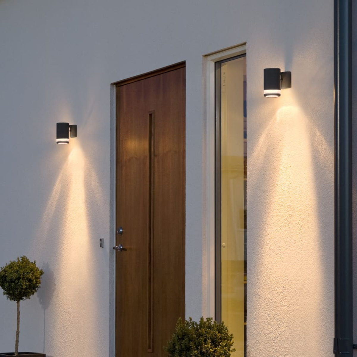 Zwei schwarze Außenwandleuchten beleuchten eine weiße Wand neben einer braunen Tür.
