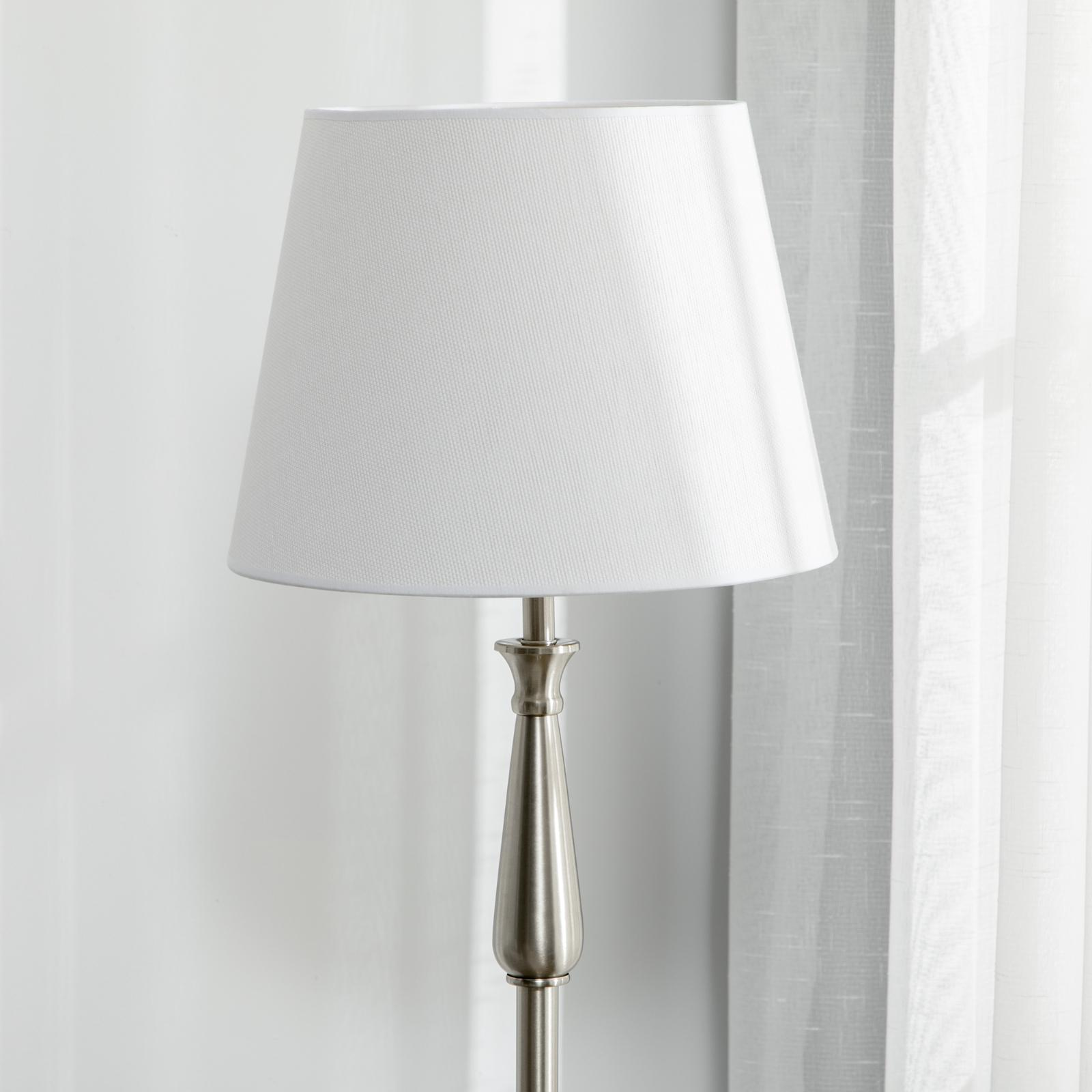Weiße Stehlampe mit weißem Lampenschirm. Der Hintergrund ist ein helles Fenster mit weißem Vorhang.