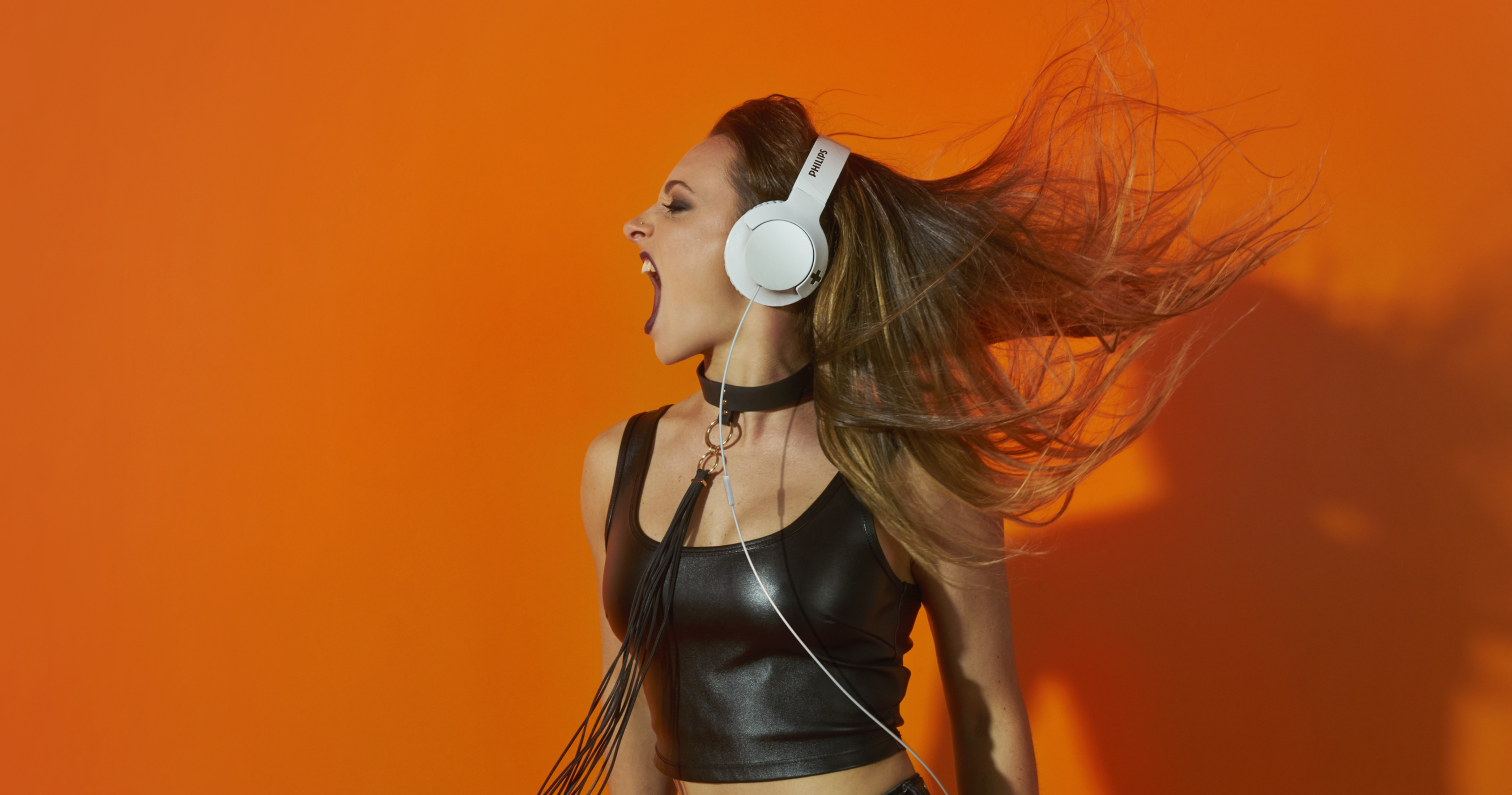 Mujer con auriculares gritando, fondo naranja. Lleva top negro y gargantilla.