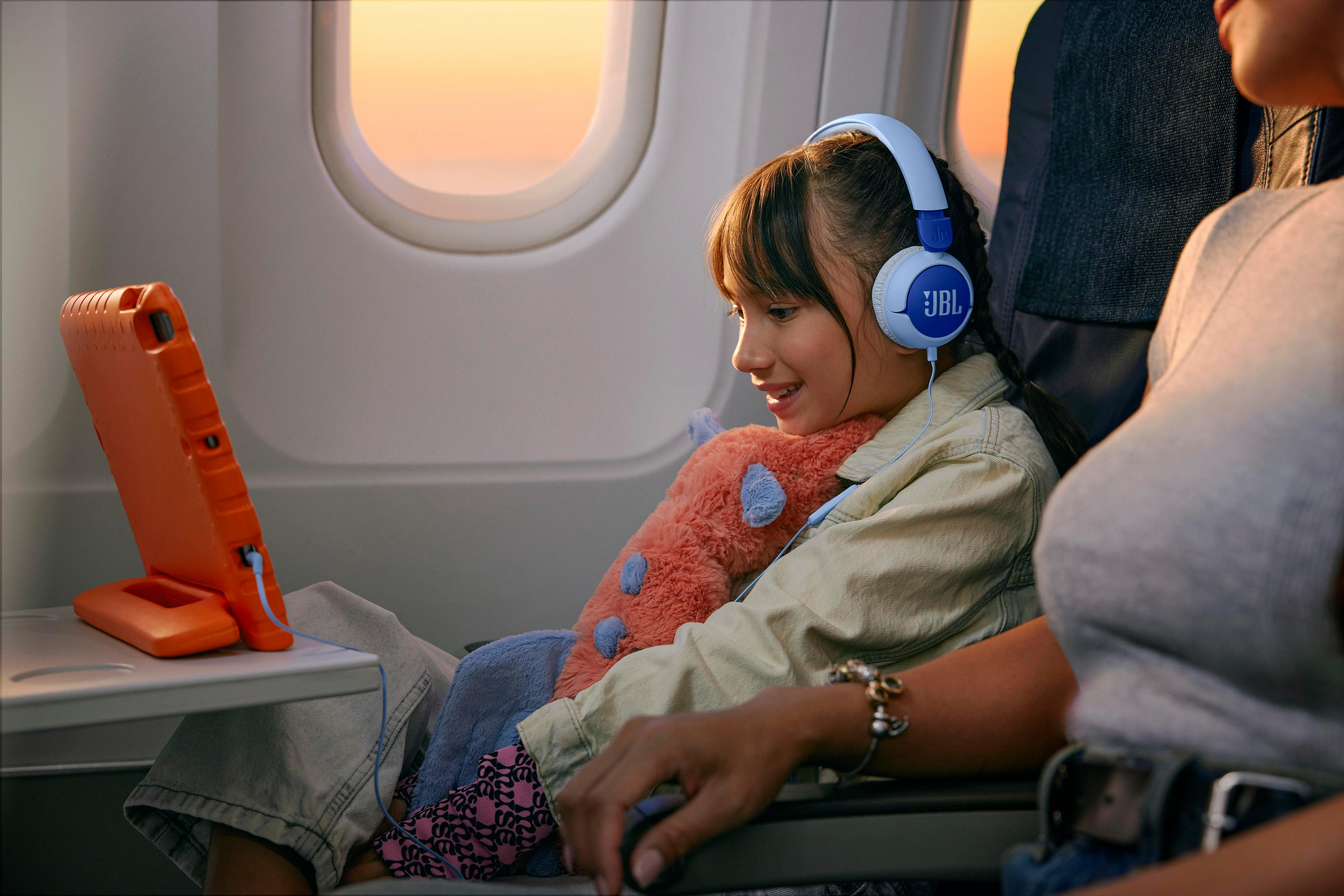 Una bambina su un aereo indossa le cuffie e guarda un tablet. Una donna le siede accanto, tenendo un giocattolo.