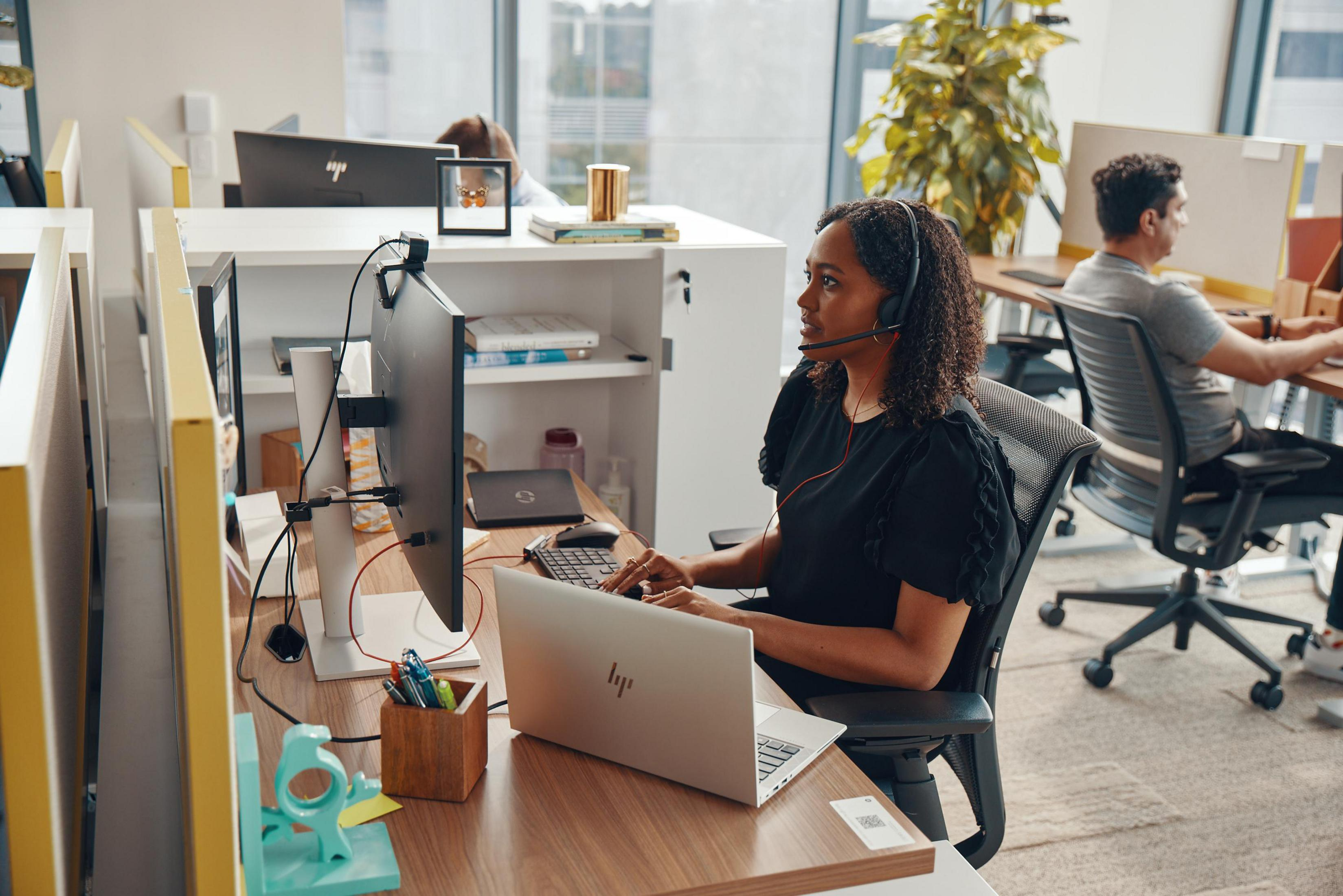 Kobieta w zestawie słuchawkowym przy biurku. Jest w środowisku biurowym.
