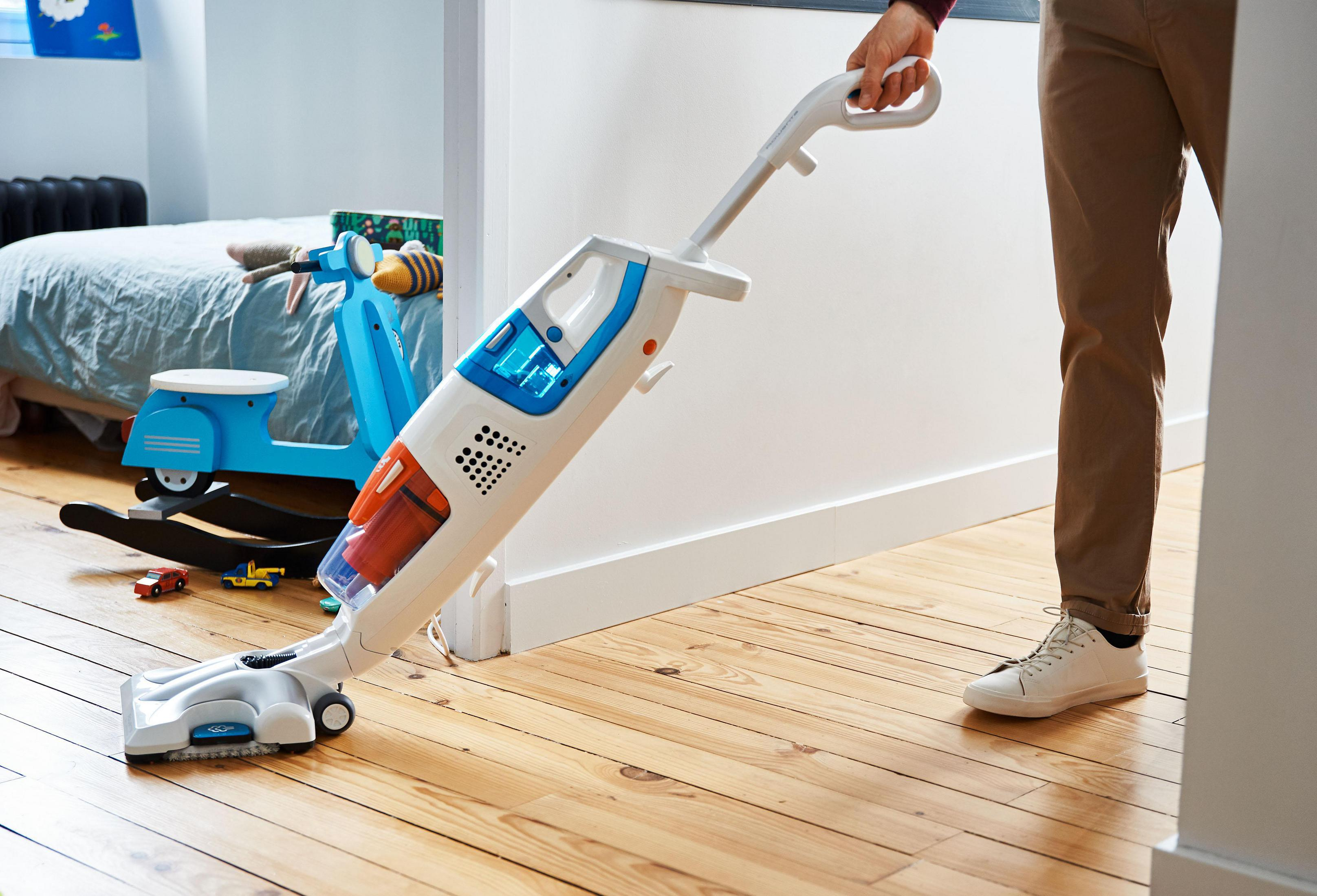 Una persona aspira una habitación con una aspiradora blanca y azul. Un scooter y juguetes en el fondo.