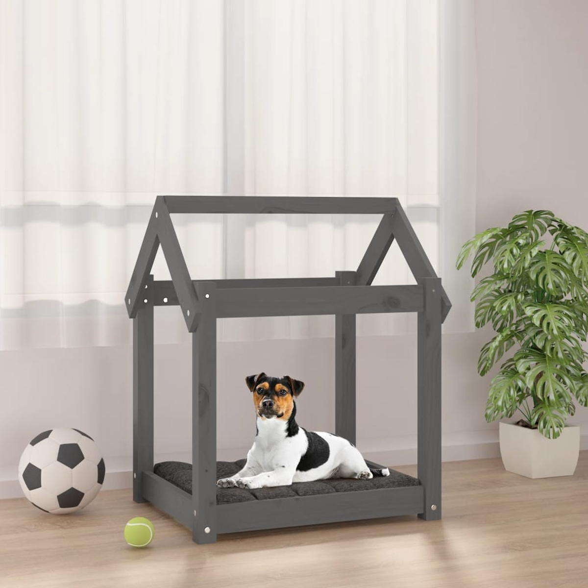 Ein kleiner Hund ruht in einem grauen Hundehäuschen aus Holz, mit einem Ball und einer Pflanze in der Nähe.