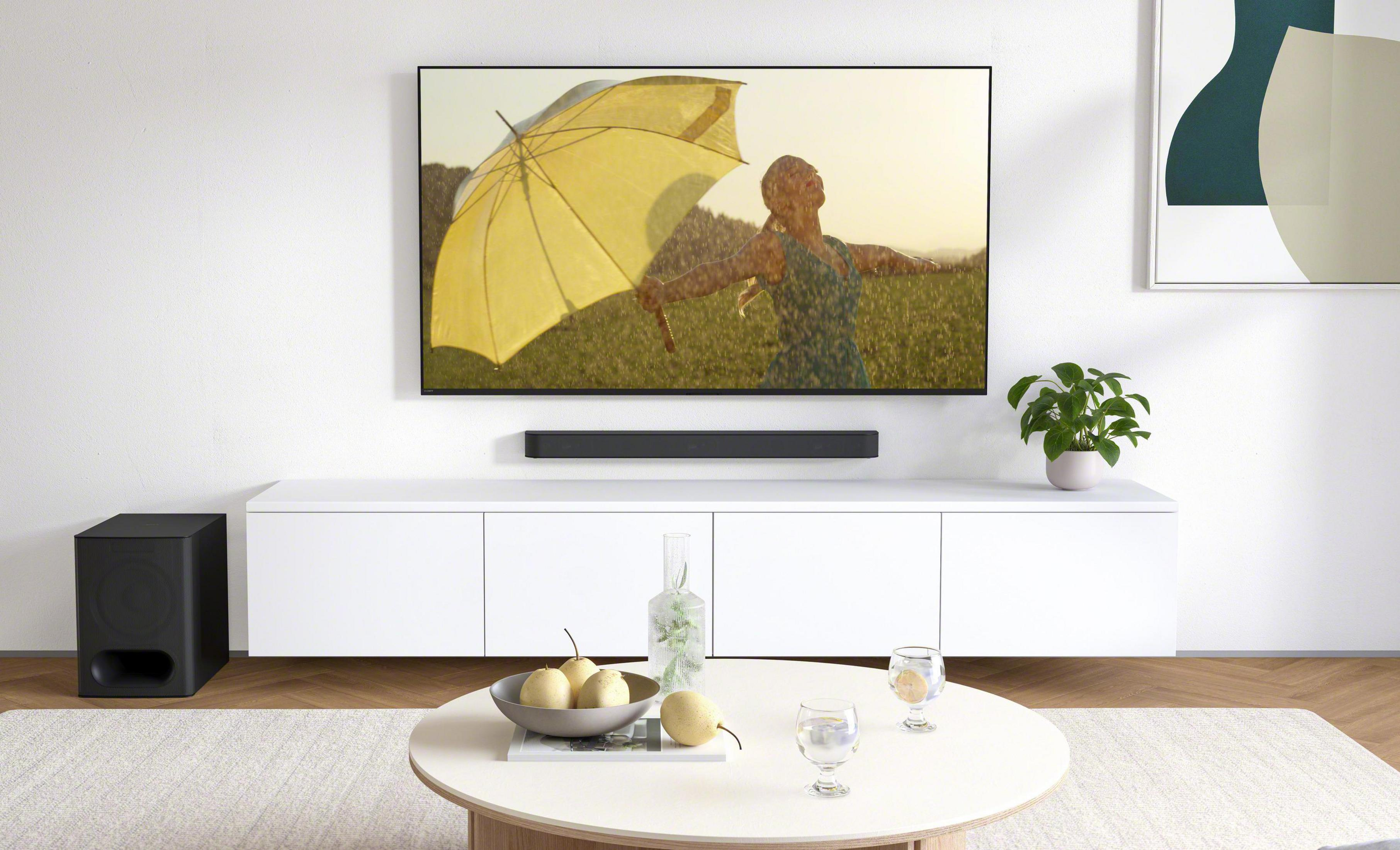 Telewizor z kobietą z parasolem. Soundbar poniżej. Biała szafka, stolik kawowy z dekoracjami.