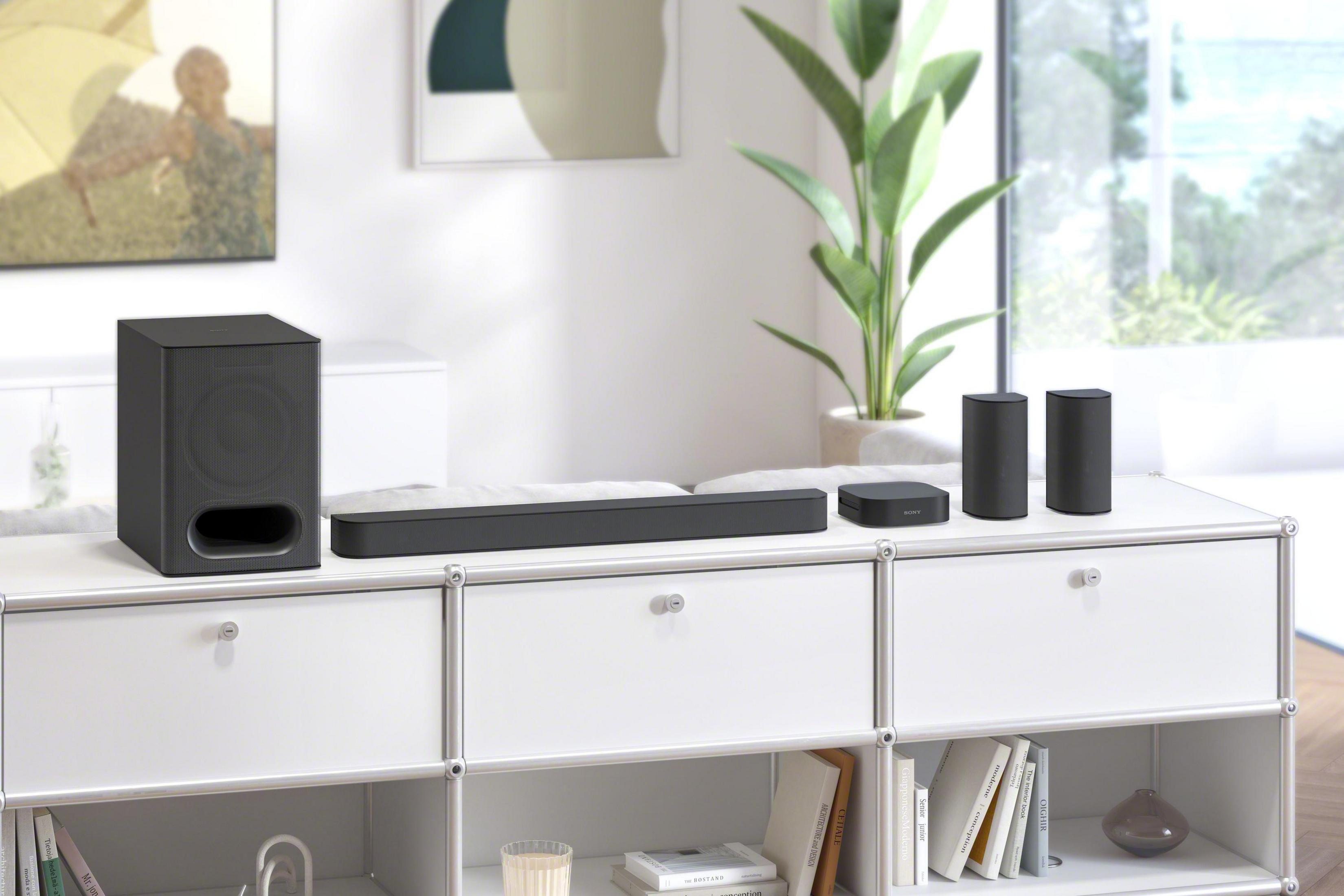 Czarny soundbar, subwoofer i dwa głośniki na białej szafce z książkami i wazonem, z tyłu zielona roślina.