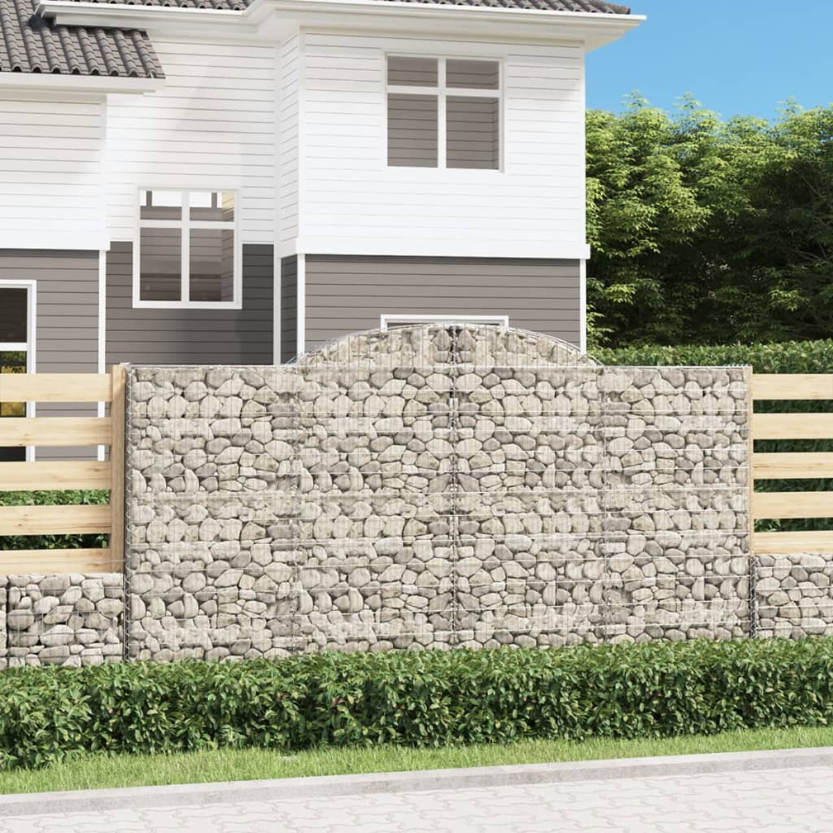Gabionenwand als Gartenzaun. Mit Steinen gefülltes Drahtgeflecht, mit Holzzaun und Haus im Hintergrund.