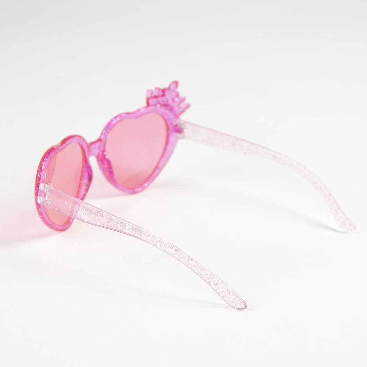 Gafas de sol rosas en forma de corazón con detalle de corona, en ángulo sobre una superficie blanca.