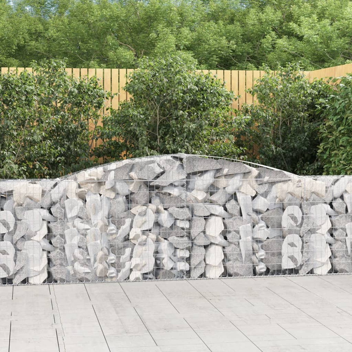 Eine Stein-Gabionenwand in einem Garten. Hinter der Wand sind grüne Sträucher und ein Holzzaun.