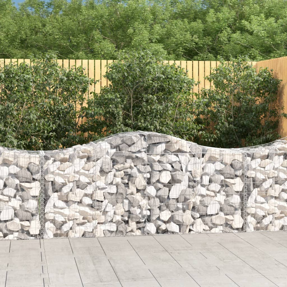 Mit Steinen gefüllte Gabionenmauer vor Holzzaun und grünem Laub.