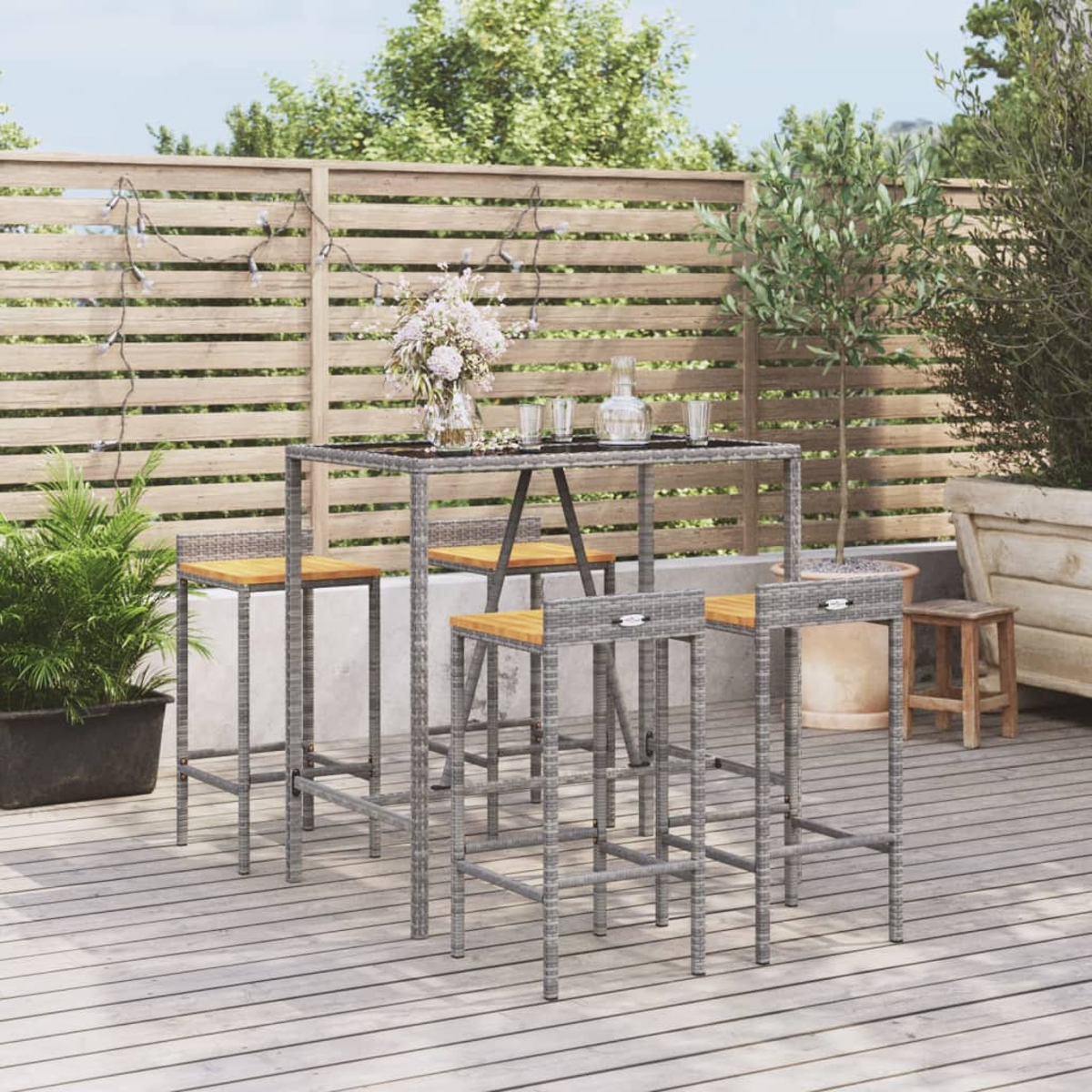 Bartisch im Freien mit vier Hockern. Der Tisch hat eine Holzplatte und ein graues Korbgittergestell. Holzterrasse im Hintergrund.