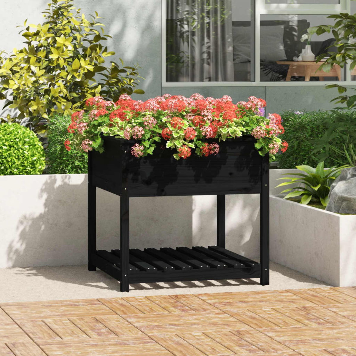 Schwarzer Pflanzkasten mit roten Blumen auf Holzterrasse. Gebäude und Pflanzen im Hintergrund.