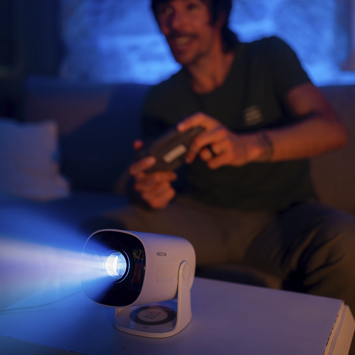 Mężczyzna uśmiecha się, trzymając kontroler do gier, podczas gdy projektor emituje niebieskie światło na stole.