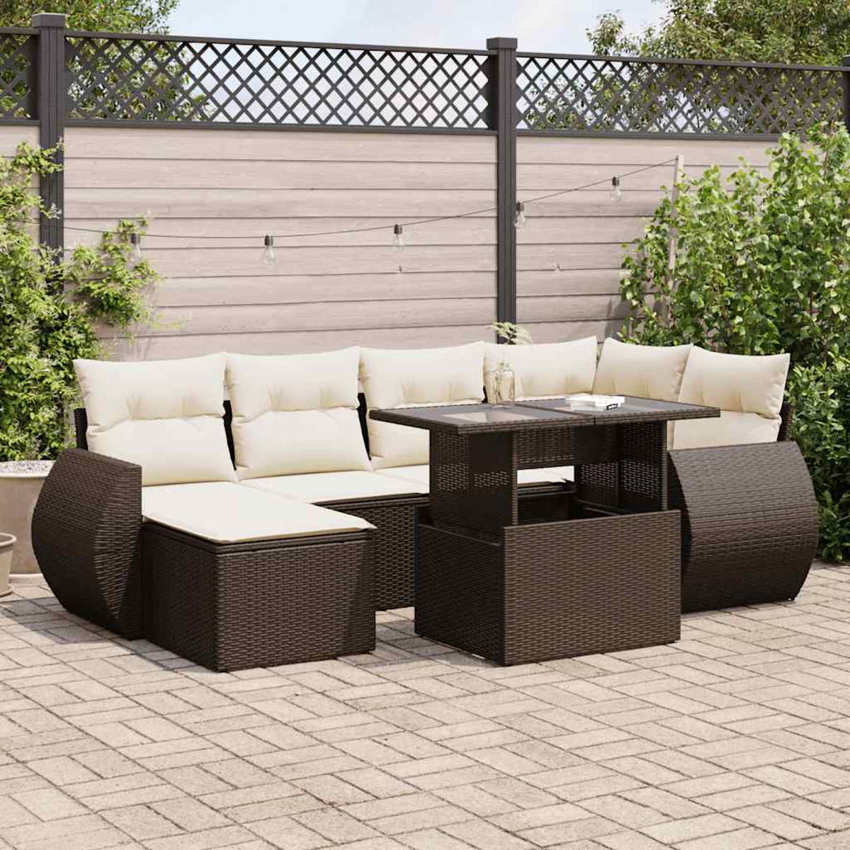 Divano componibile in vimini marrone con cuscini color crema, tavolo, su patio con recinzione e verde.
