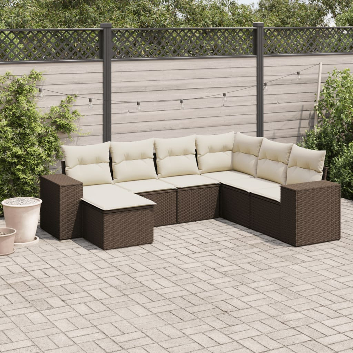 Set divano da esterno. Struttura in rattan marrone, cuscini bianchi e tavolo in legno. Patio in mattoni e recinzione.