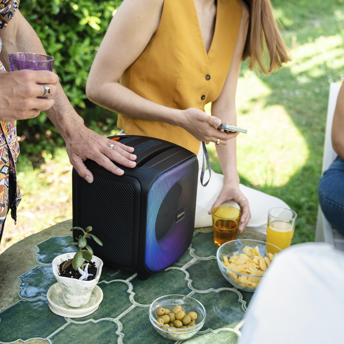 Ludzie na imprezie na świeżym powietrzu. Głośnik i przekąski na stole z napojami. Ktoś trzyma szklankę.