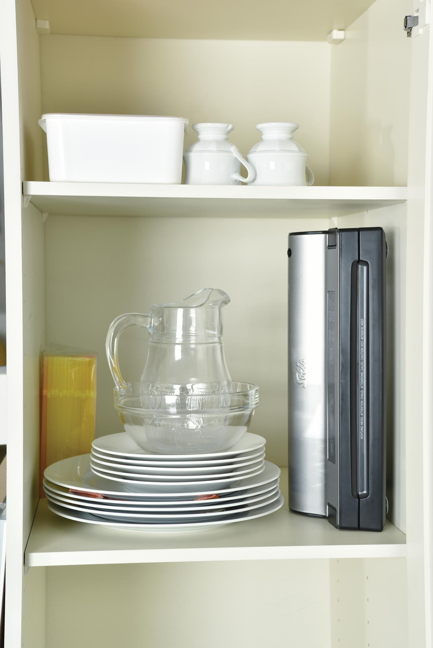 Półka z białymi pojemnikami, szklanymi naczyniami i talerzami. Obecny wysoki metalowy przedmiot.