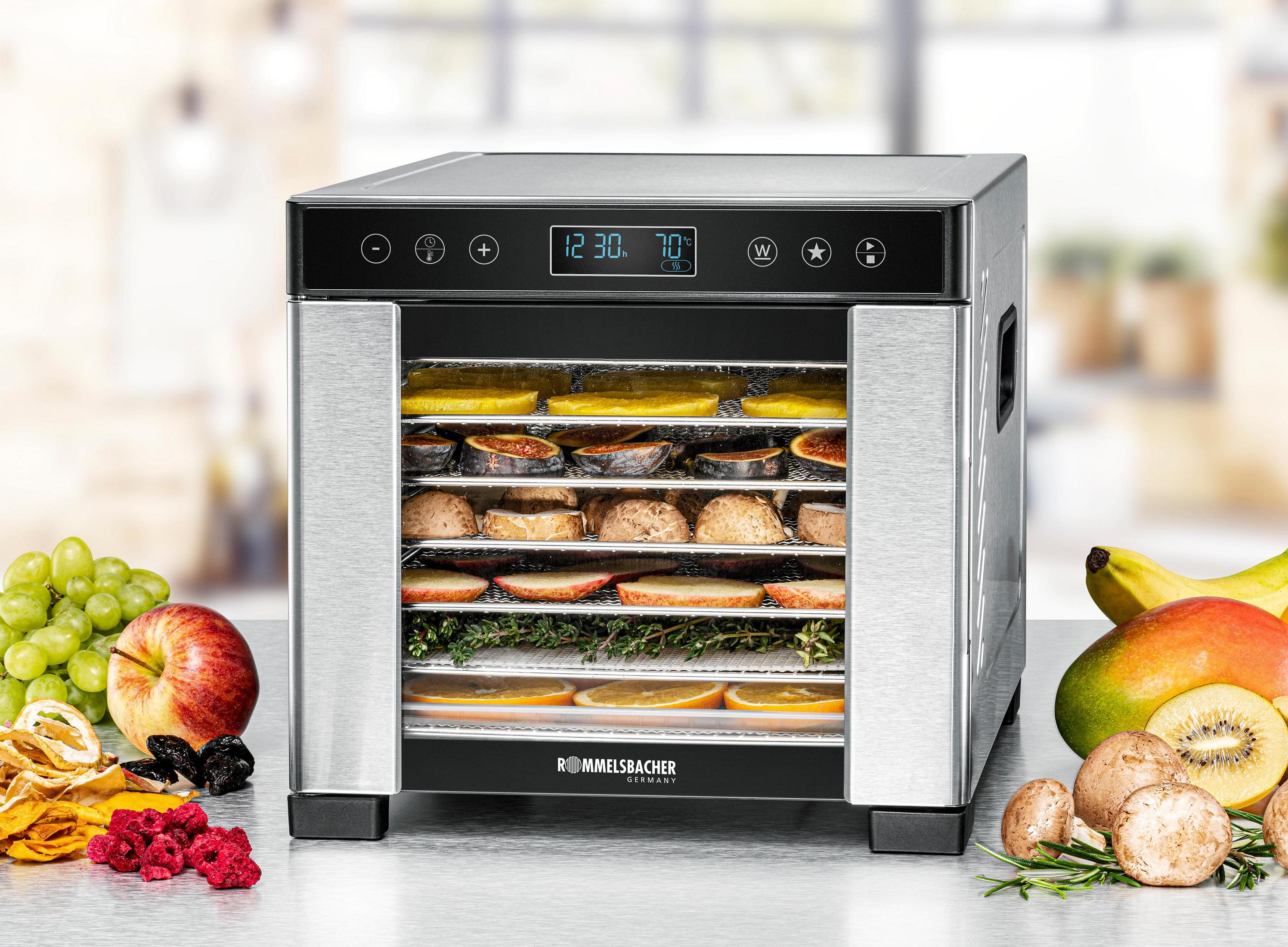 Nahrungsmittel-Dehydrator mit offener Tür, gefüllt mit verschiedenen Früchten und Kräutern. Zusätzliche Früchte auf dem Tisch.