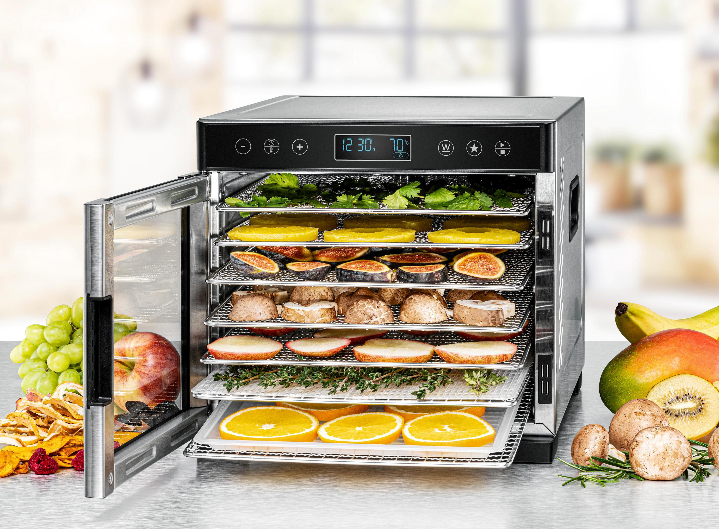 Nahrungsmittel-Dehydrator mit offener Tür, gefüllt mit verschiedenen Früchten und Kräutern. Zusätzliche Früchte auf dem Tisch.