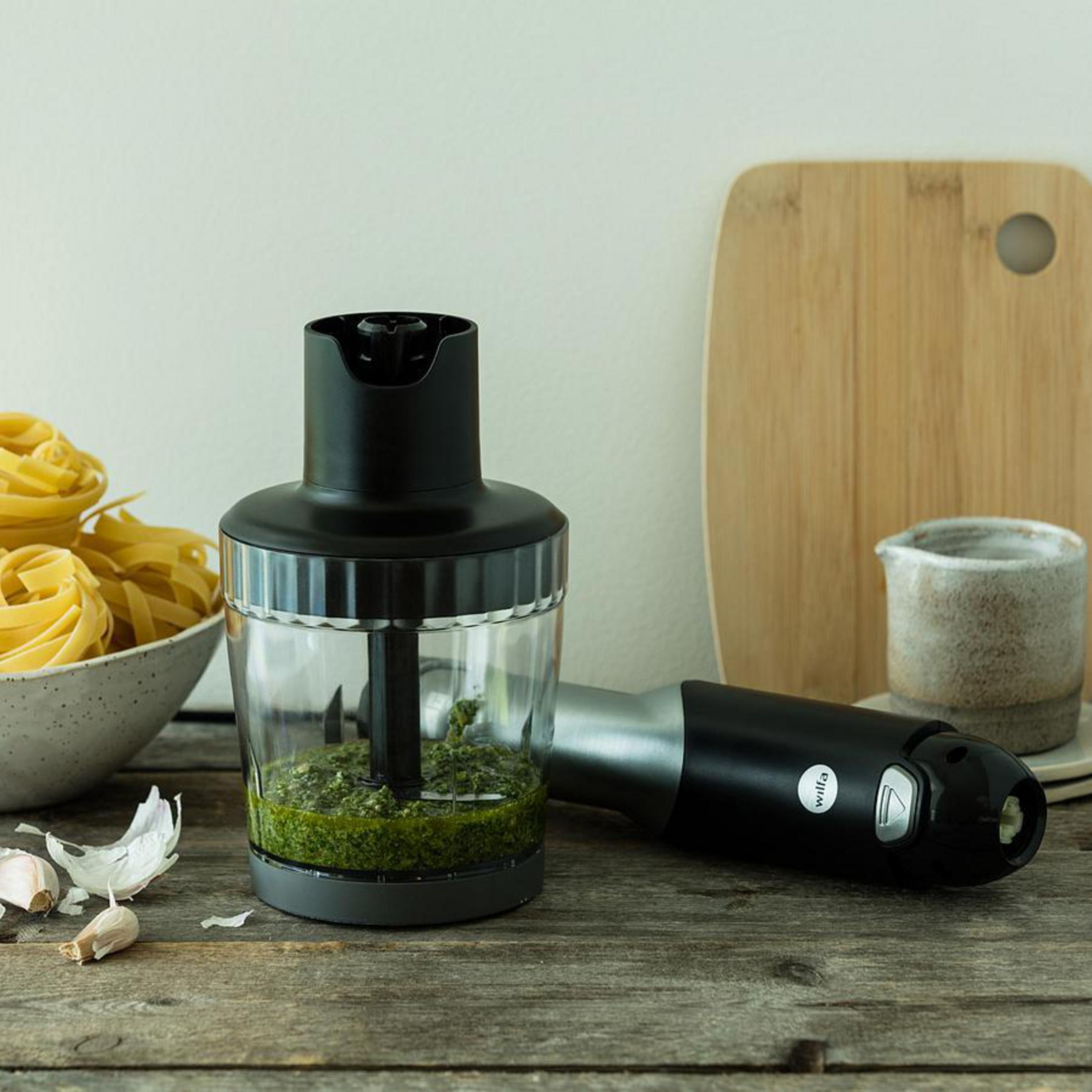 Frullatore a immersione nero e argento con pesto verde in contenitore trasparente, su legno.