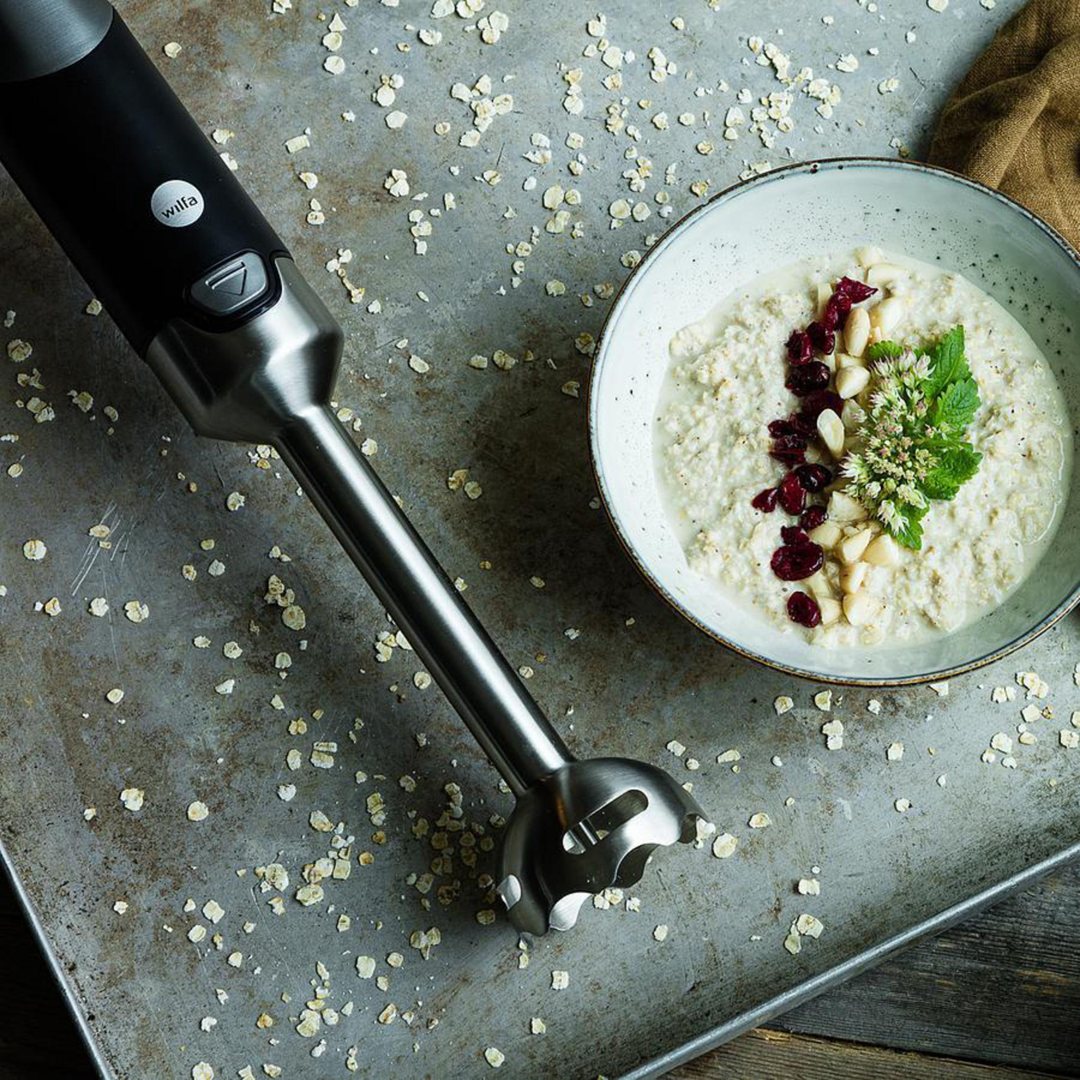 Frullatore a immersione vicino a una ciotola di farina d'avena con frutti di bosco e noci.