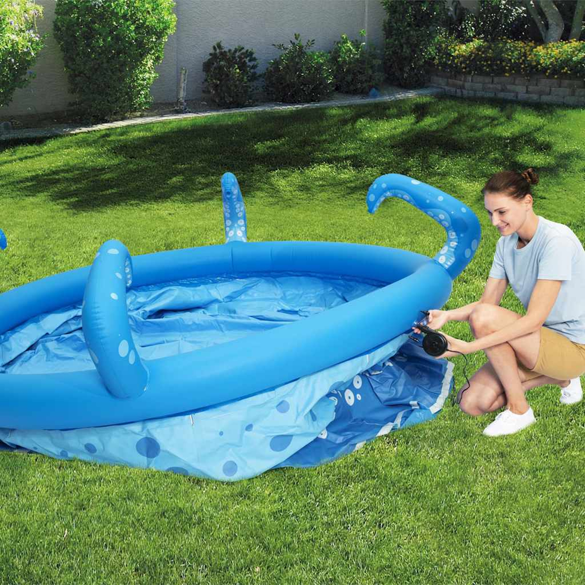 Eine Frau pumpt einen blauen, oktopusförmigen Pool auf einer grünen Wiese auf.
