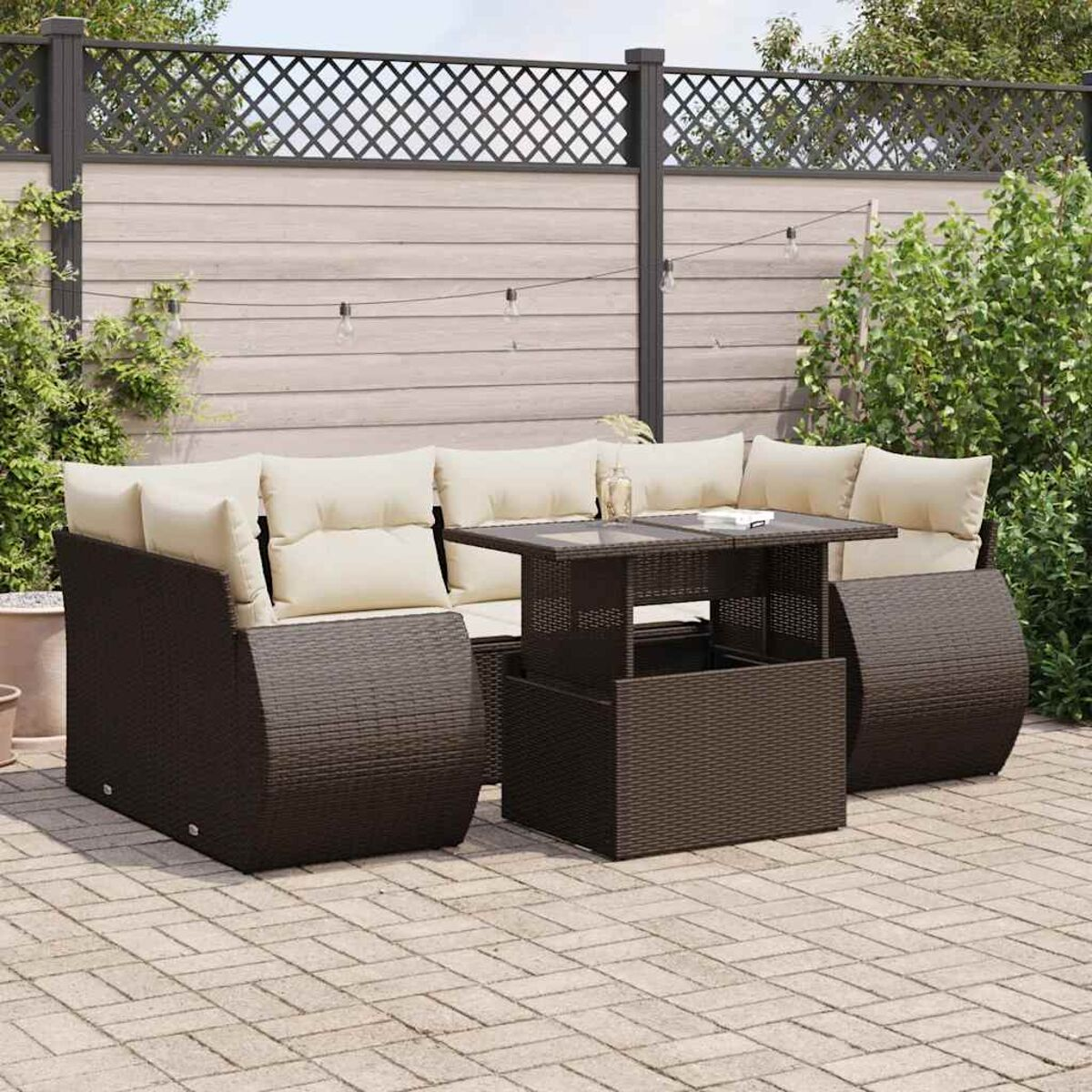 Set da patio all'aperto con mobili intrecciati marroni, cuscini color crema e un tavolo. Lo sfondo ha una recinzione e piante.