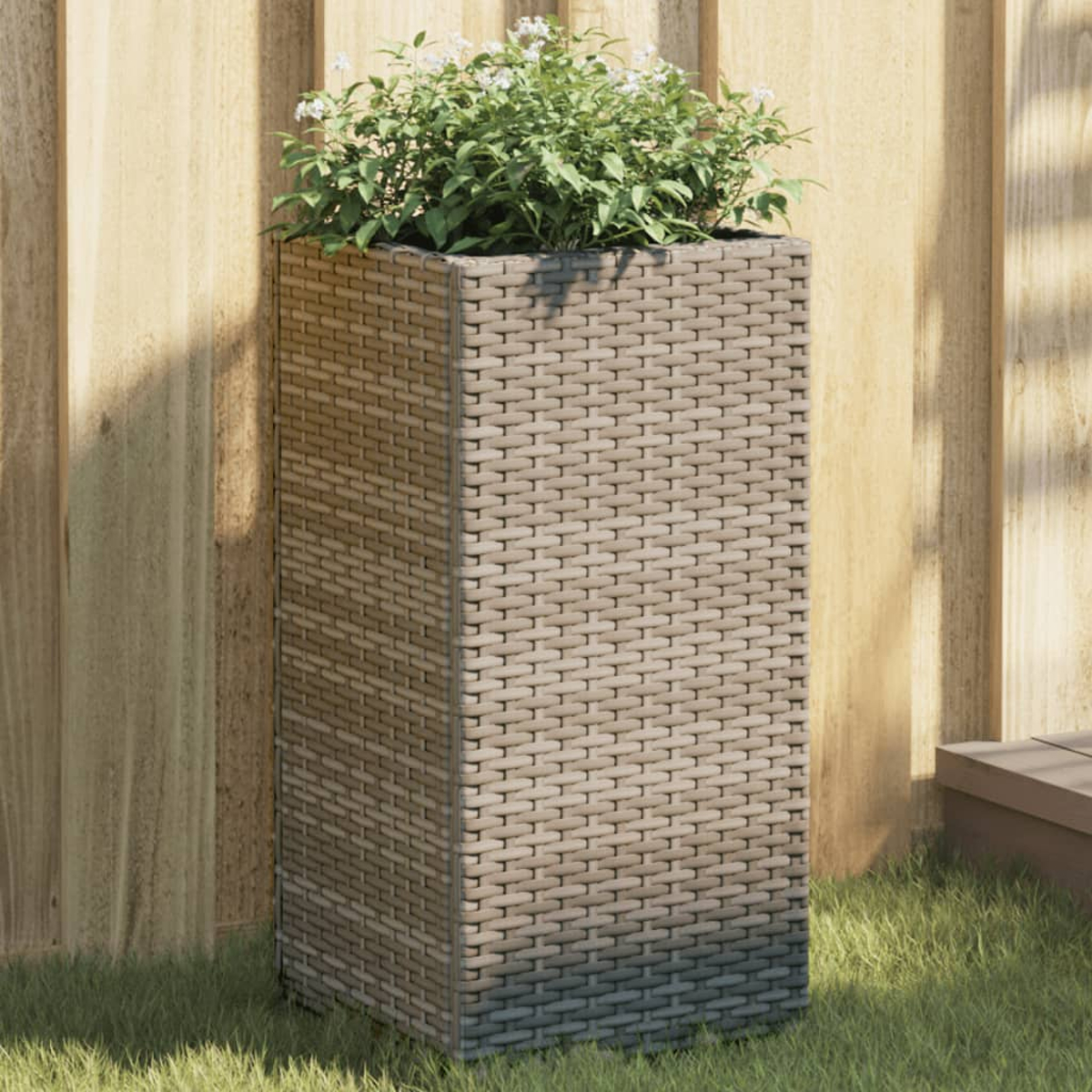 Fioriera alta in rattan grigio con una pianta. Lo sfondo è una recinzione di legno.