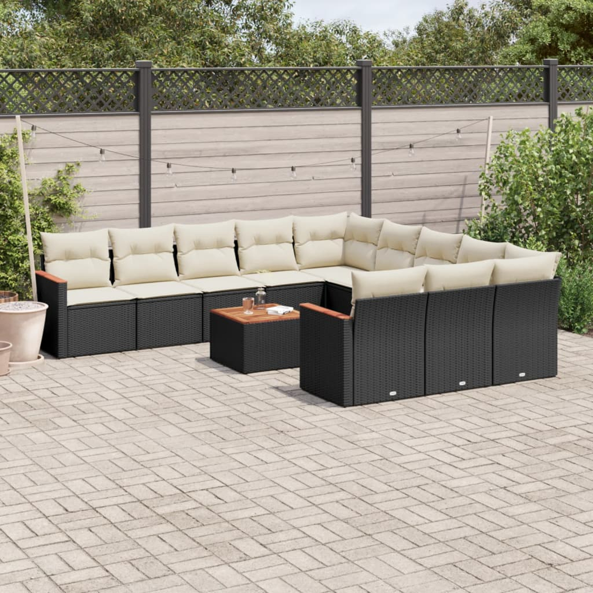 Ein schwarzes Outdoor-Sitzmöbel-Set aus Korbgeflecht mit beigen Kissen, einem Holztisch und einer gepflasterten Terrasse. Der Hintergrund ist ein Zaun.