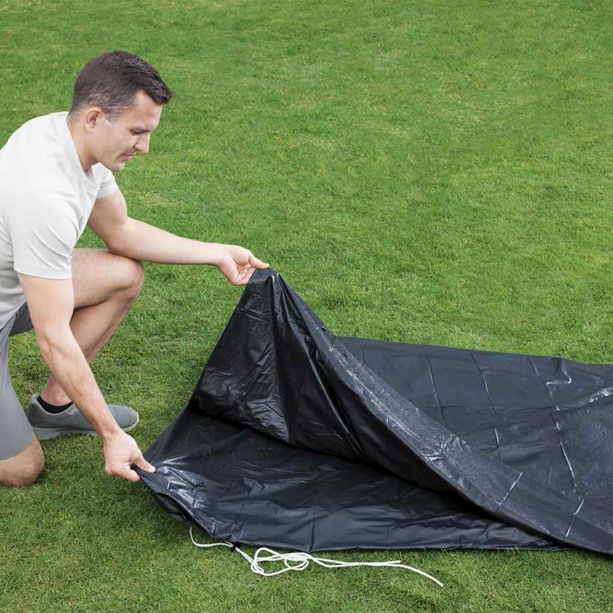 Ein Mann kniet auf grünem Gras und faltet eine schwarze Plane. Weiße Schnur ist sichtbar.