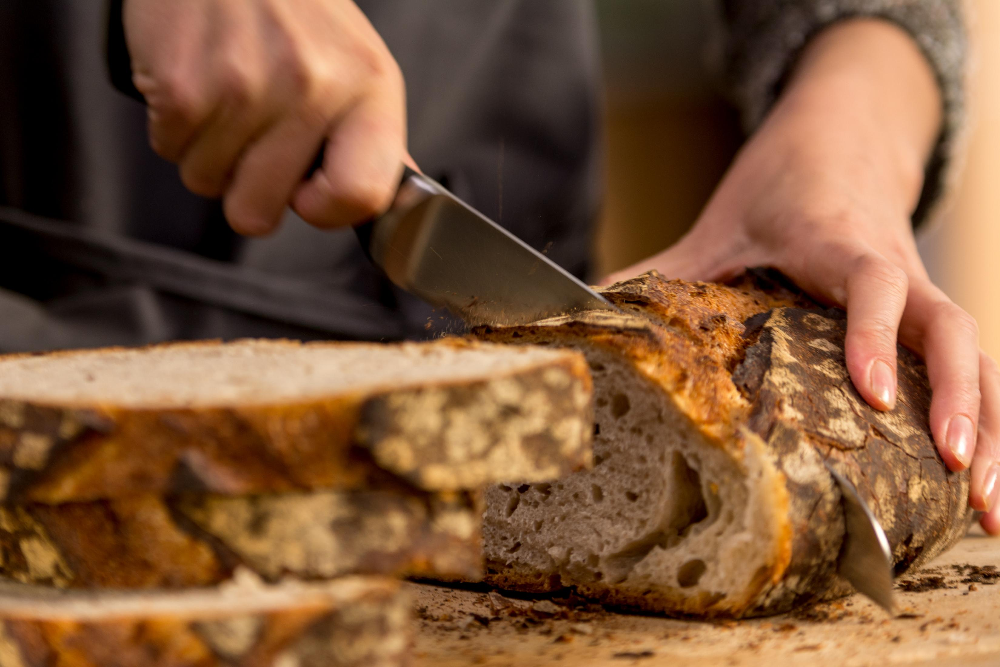 Ręce krojące chleb na drewnianej desce do krojenia nożem z ząbkami. Chleb jest pokrojony.
