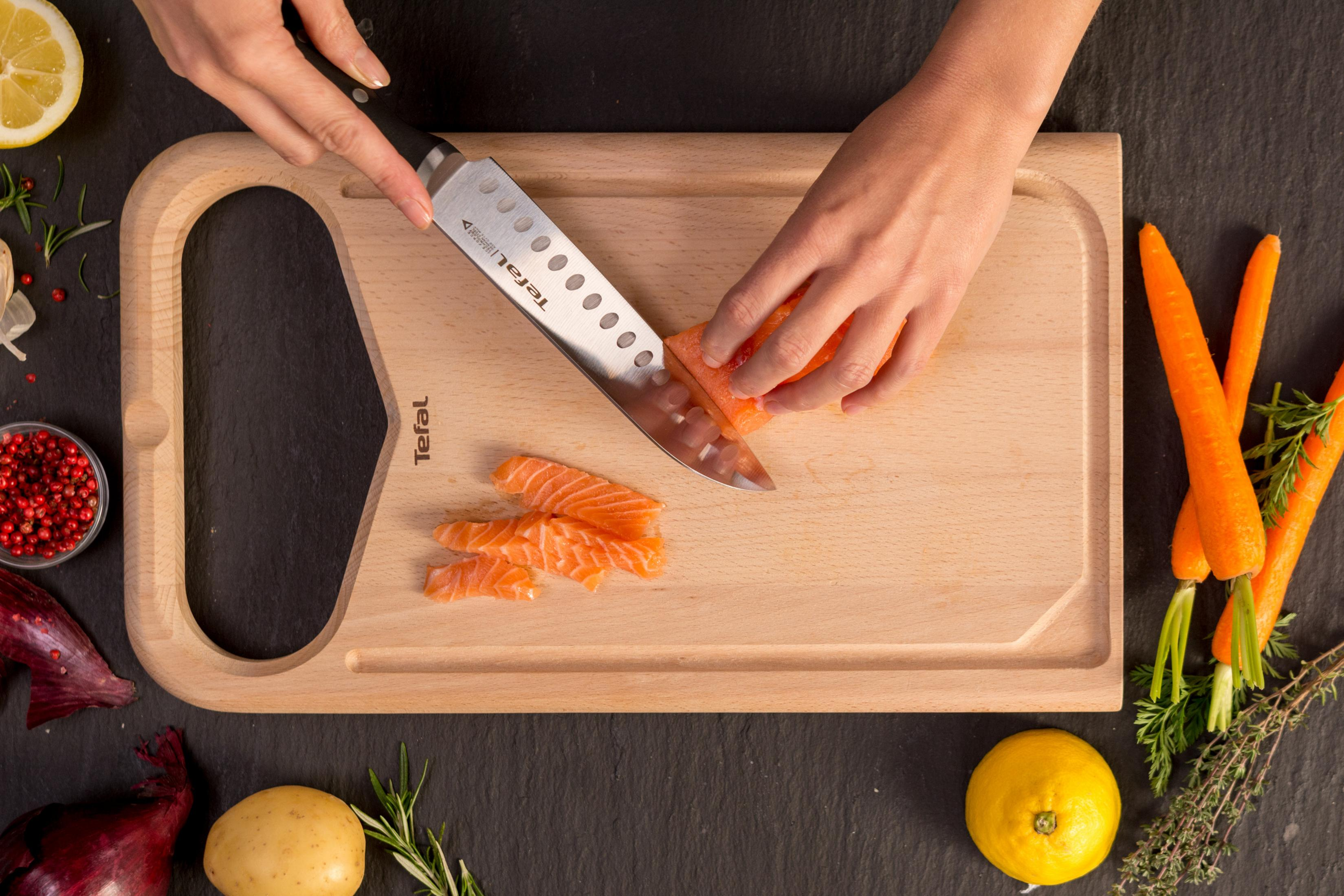 Eine Person schneidet Lachs mit einem Tefal-Messer auf einem Holzschneidebrett mit anderen Zutaten.