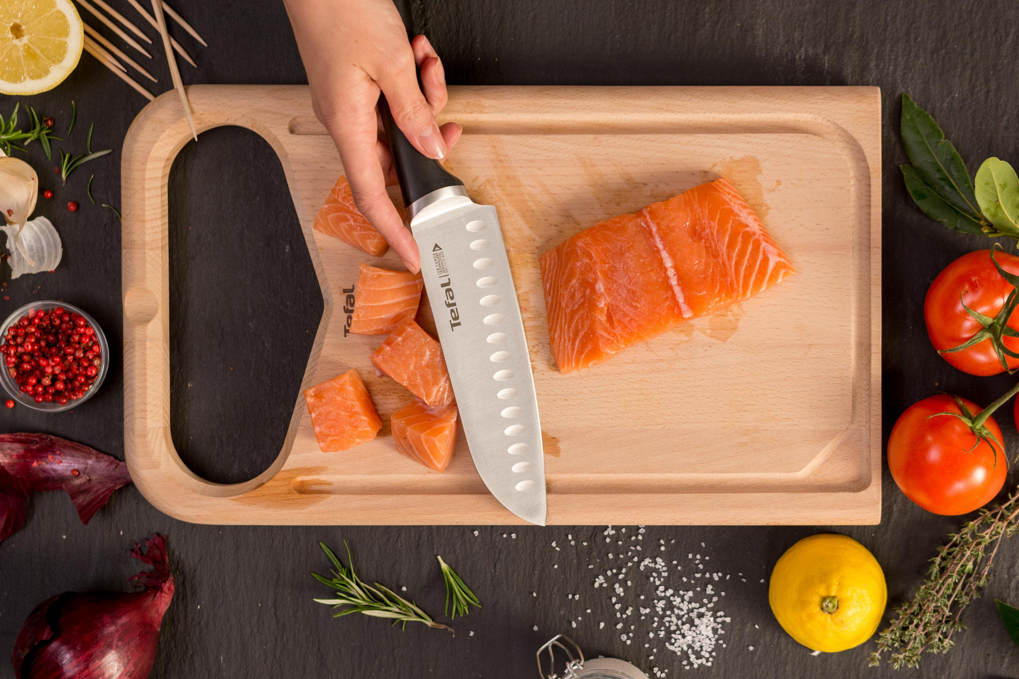 Eine Hand hält ein Messer und schneidet Lachs auf einem Holzbrett mit Zutaten.