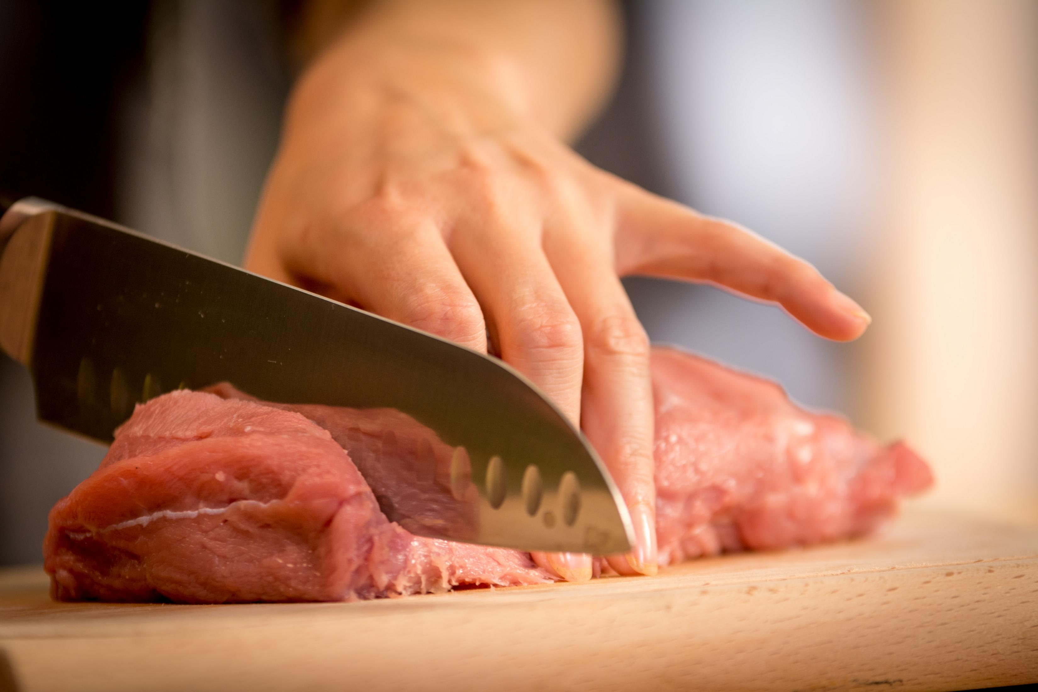 Eine Hand schneidet mit einem Messer rohes Fleisch auf einem Holzschneidebrett.