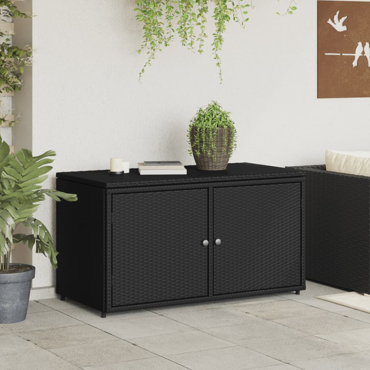 Schwarzer Rattan-Aufbewahrungsschrank für den Außenbereich mit flacher Oberseite. Eine Pflanze und eine Kerze stehen oben.