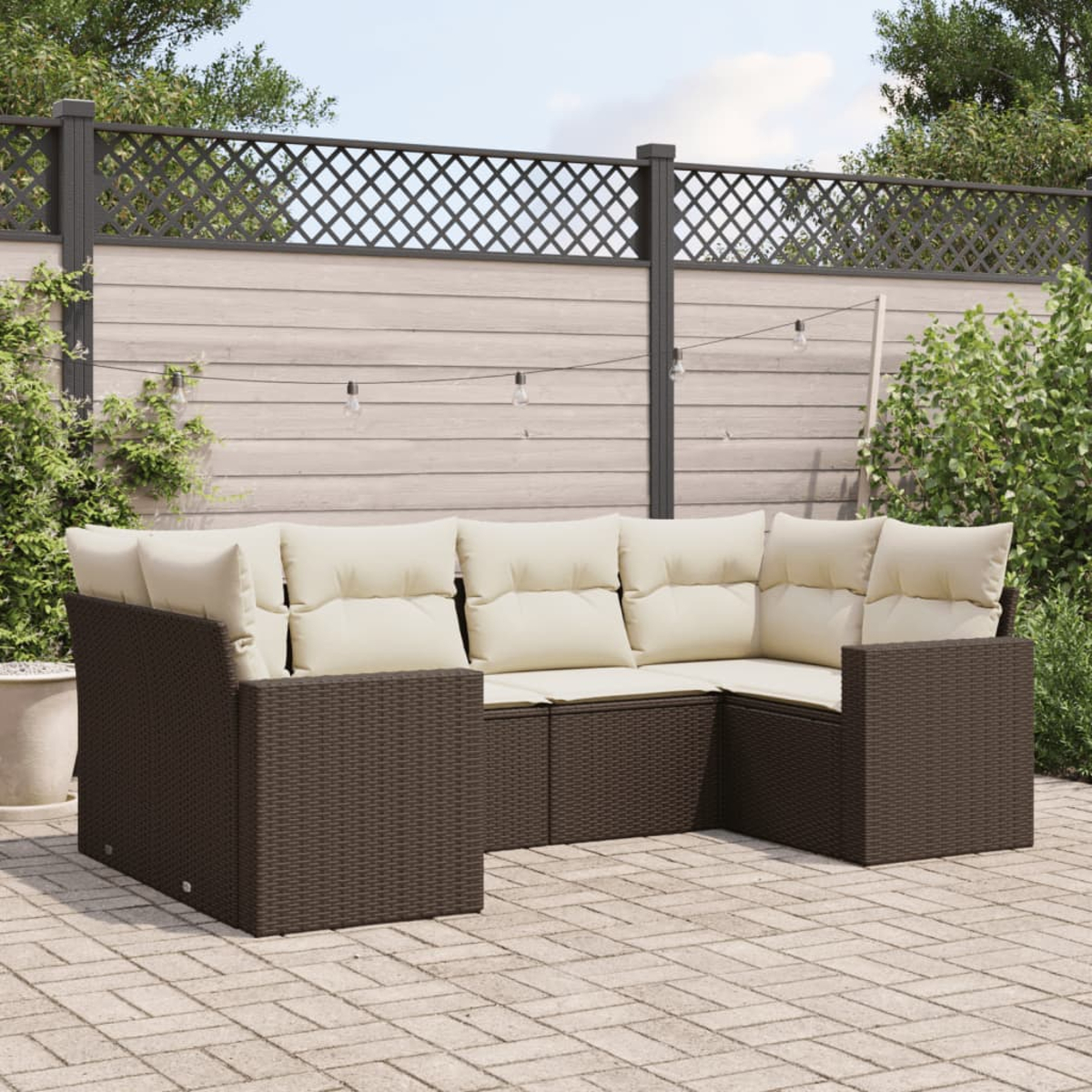 Ein braunes Ecksofa aus Korbgeflecht mit cremefarbenen Kissen auf einer Ziegelterrasse, mit Zaun und Laub.