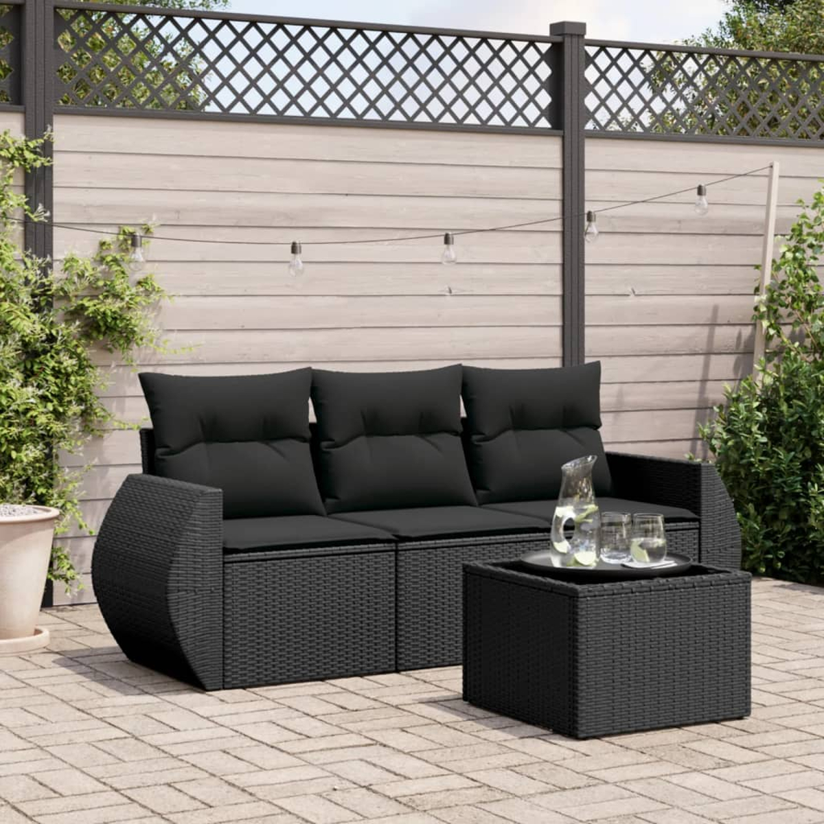 Schwarzes Outdoor-Wicker-Sofaset mit Kissen, Tisch und Lichterketten im Hintergrund.