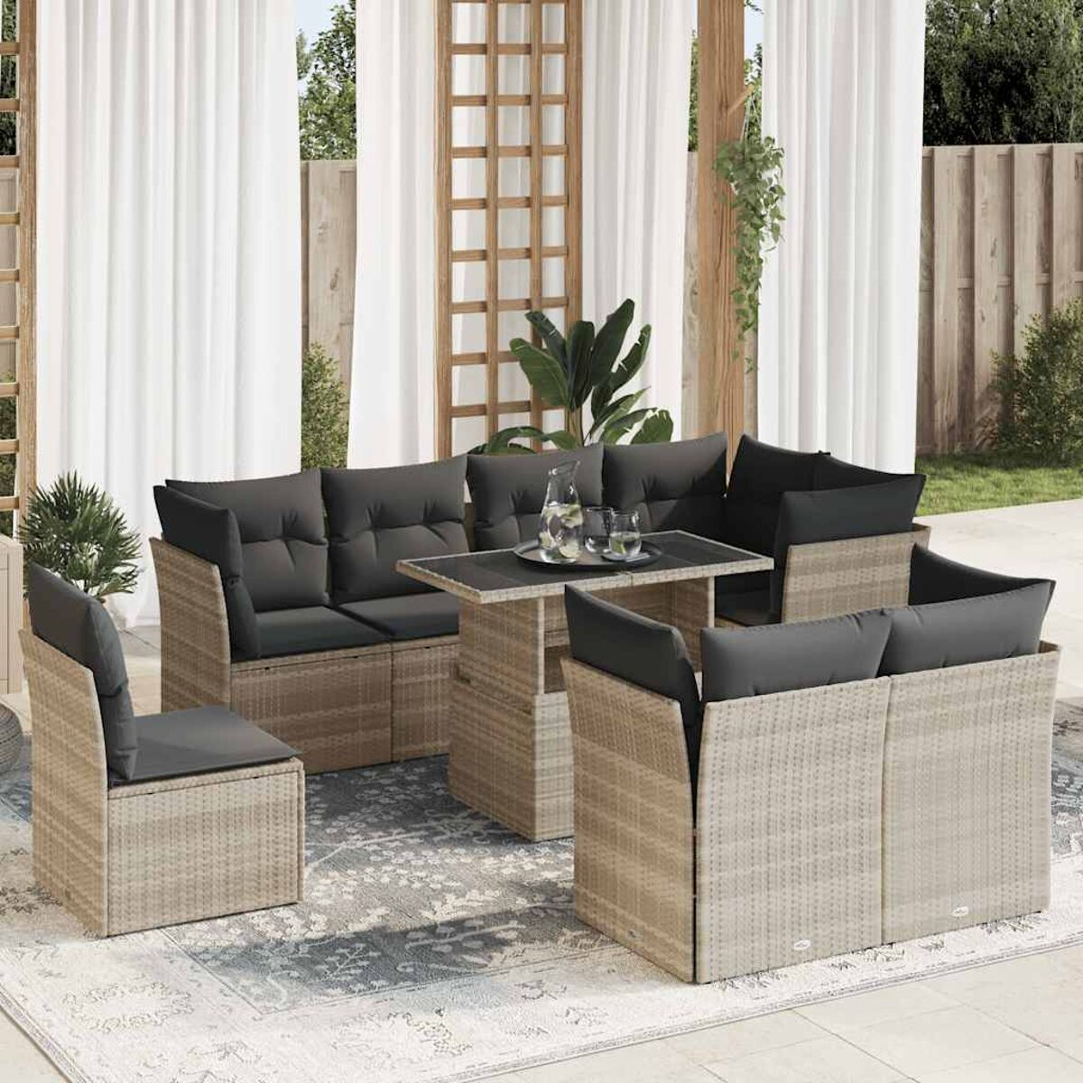 Set di mobili in rattan per esterni con divano, sedie e tavolo. Tende bianche e vegetazione.
