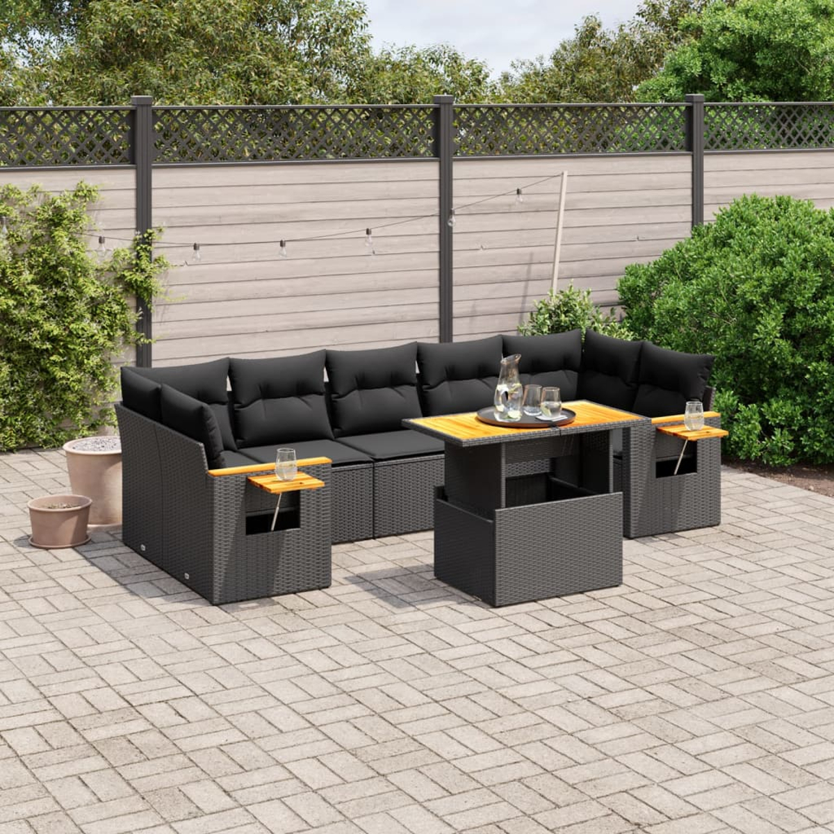 Ein Terrassenset mit schwarzen Kissen und einem Holztisch. Das Set befindet sich auf einer Backsteinterrasse mit Pflanzen.