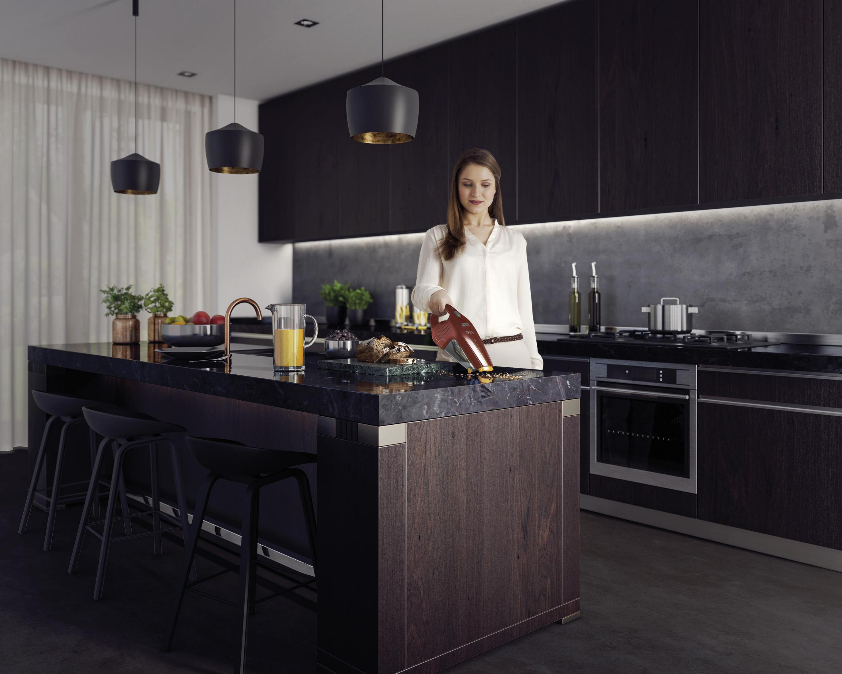 Mujer en la cocina con una aspiradora. Se ven gabinetes oscuros, isla y electrodomésticos.