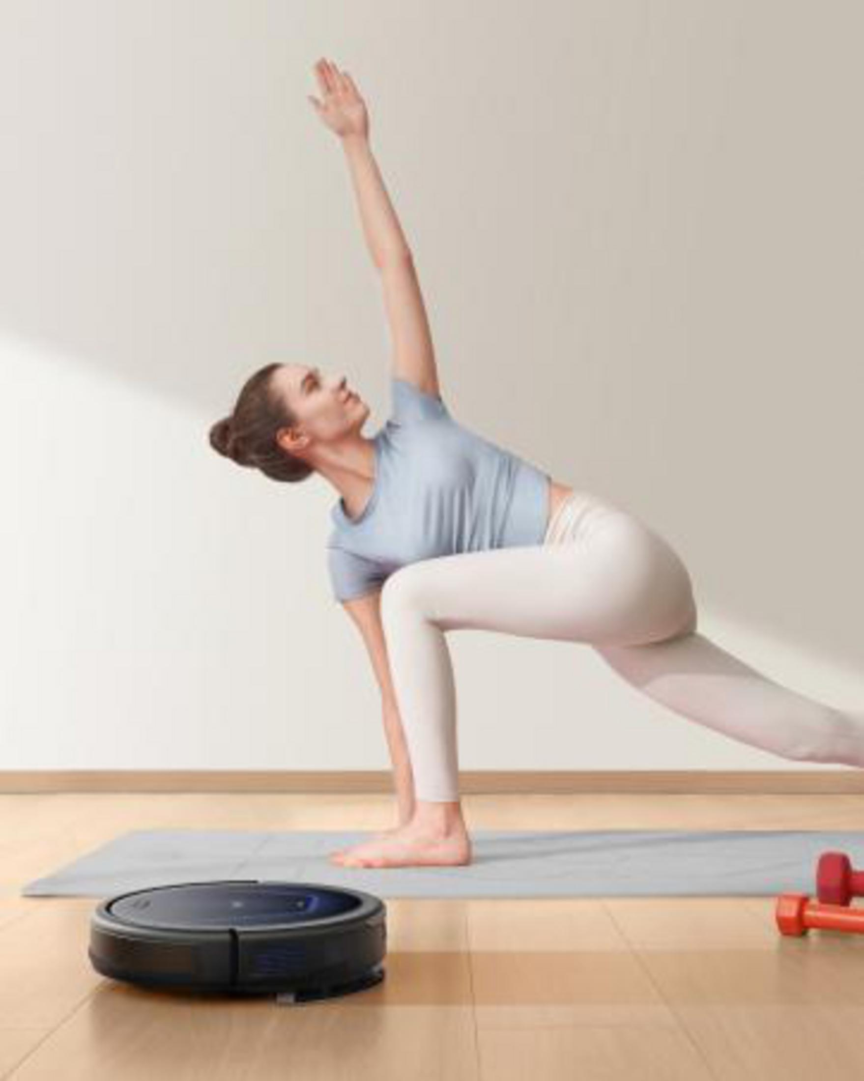 Kobieta uprawiająca jogę, robot sprzątający na podłodze i hantle w pobliżu.