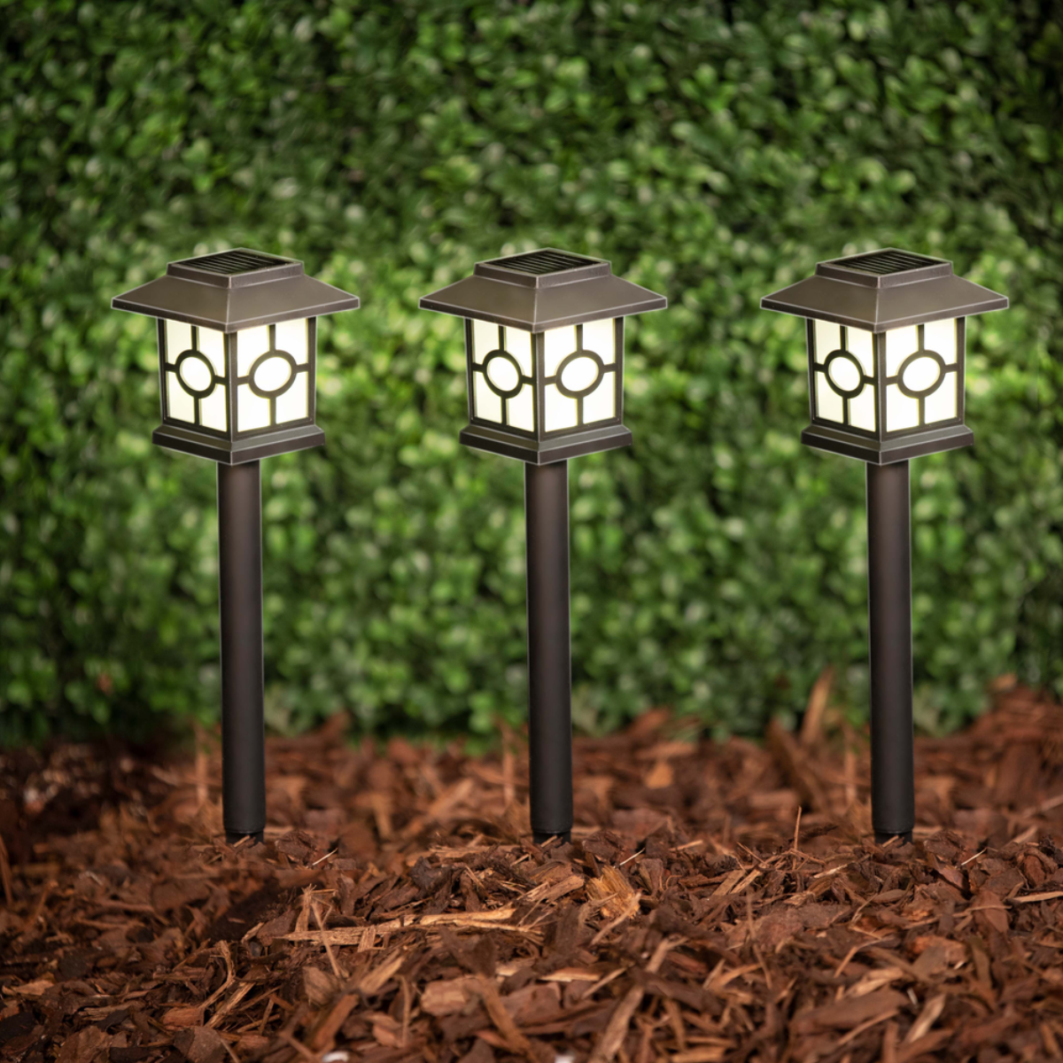 Three solar garden lights with square tops and dark posts in wood chips.