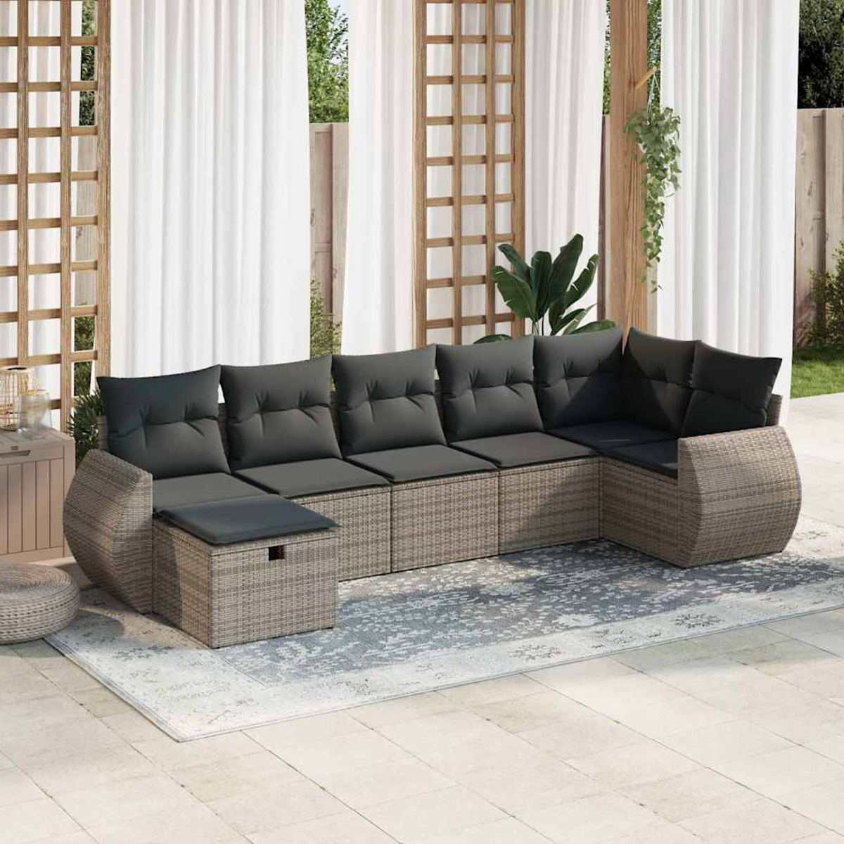 Un sofá seccional de mimbre gris con cojines negros, sobre una alfombra. Cortinas blancas al fondo. Plantas visibles.