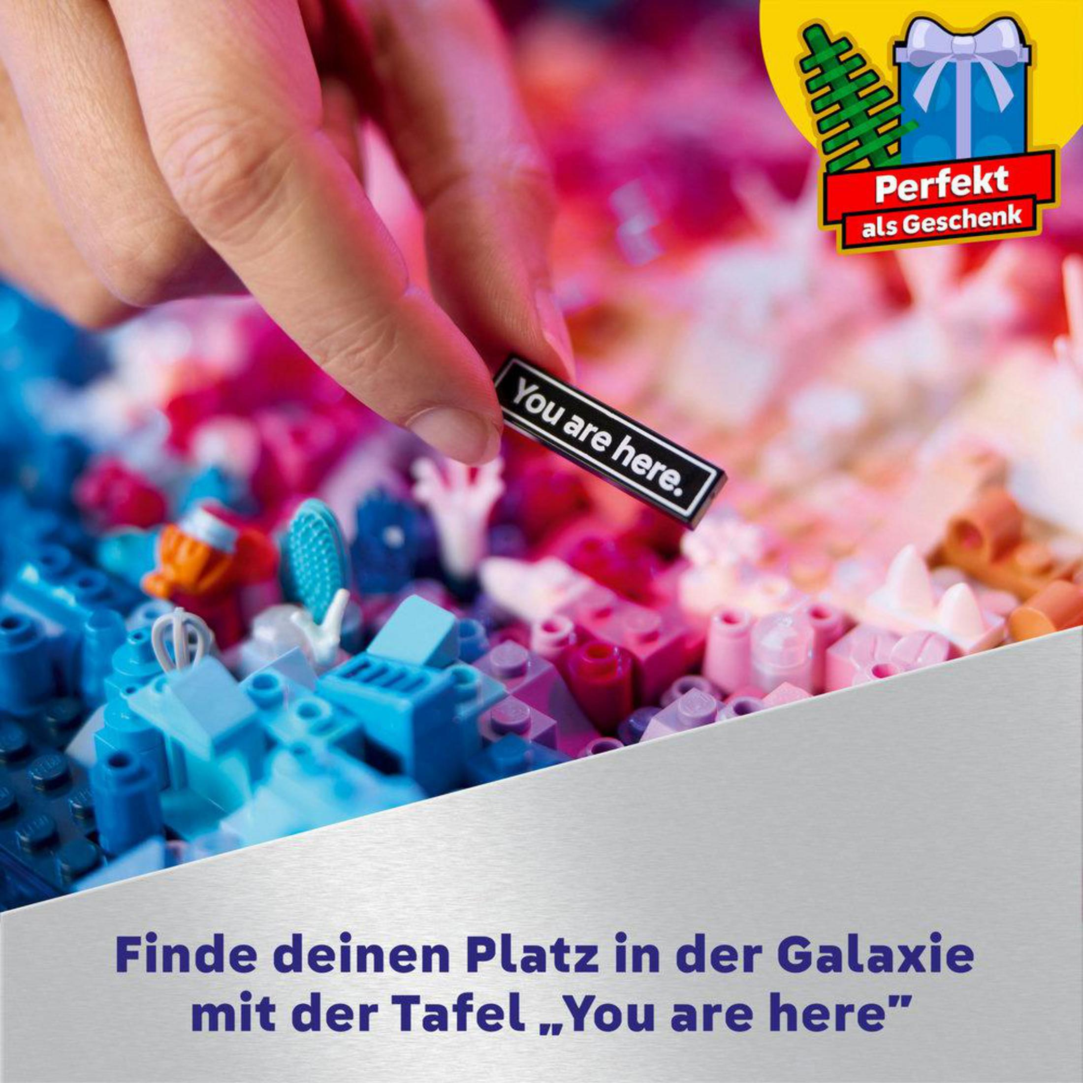 Eine Hand, die eine LEGO-Kachel hält, auf der steht: „You are here“. Neben einem bunten LEGO-Galaxiebau.
