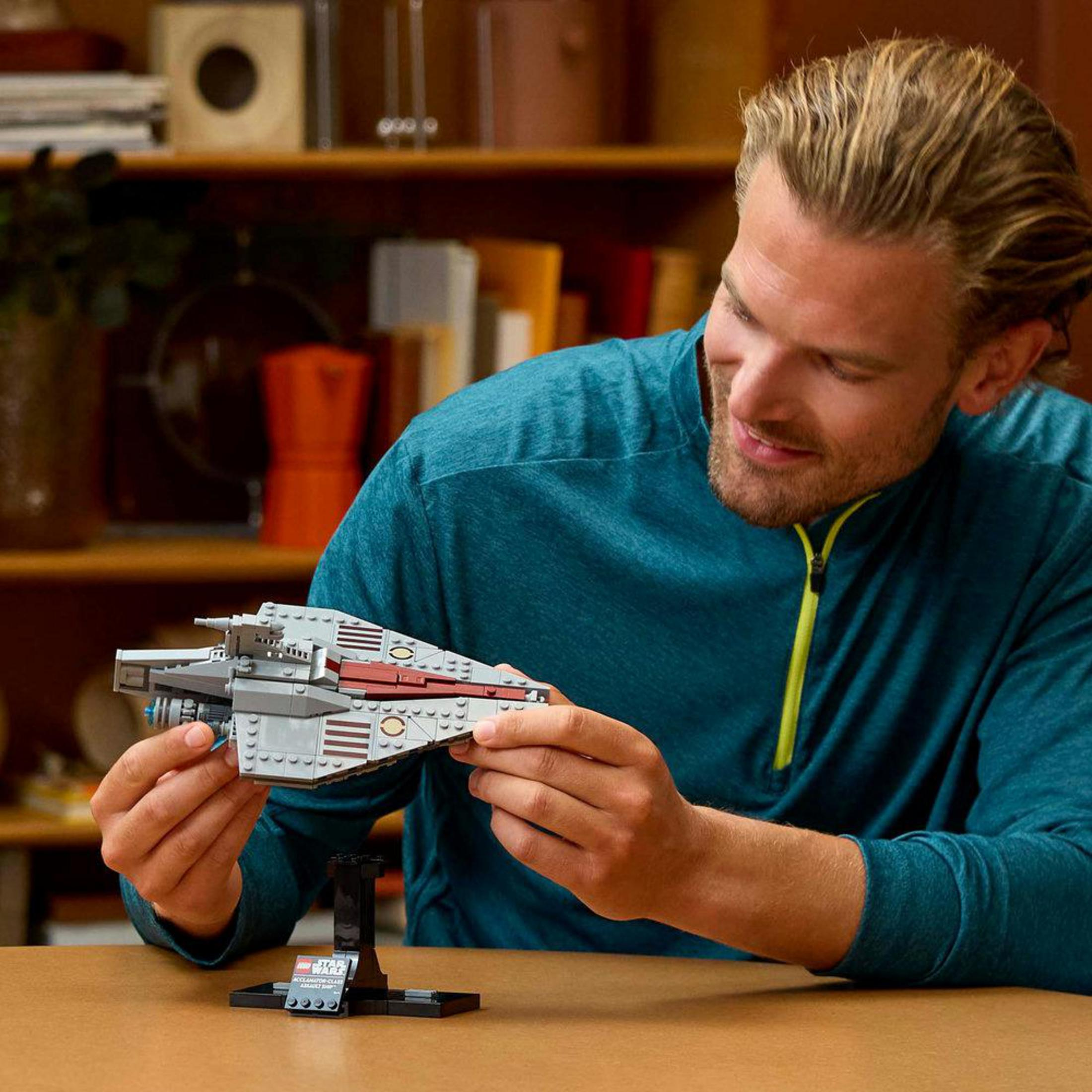 Blondyn trzymający model statku LEGO Star Wars. Model jest jasnoszary z czerwonymi paskami.