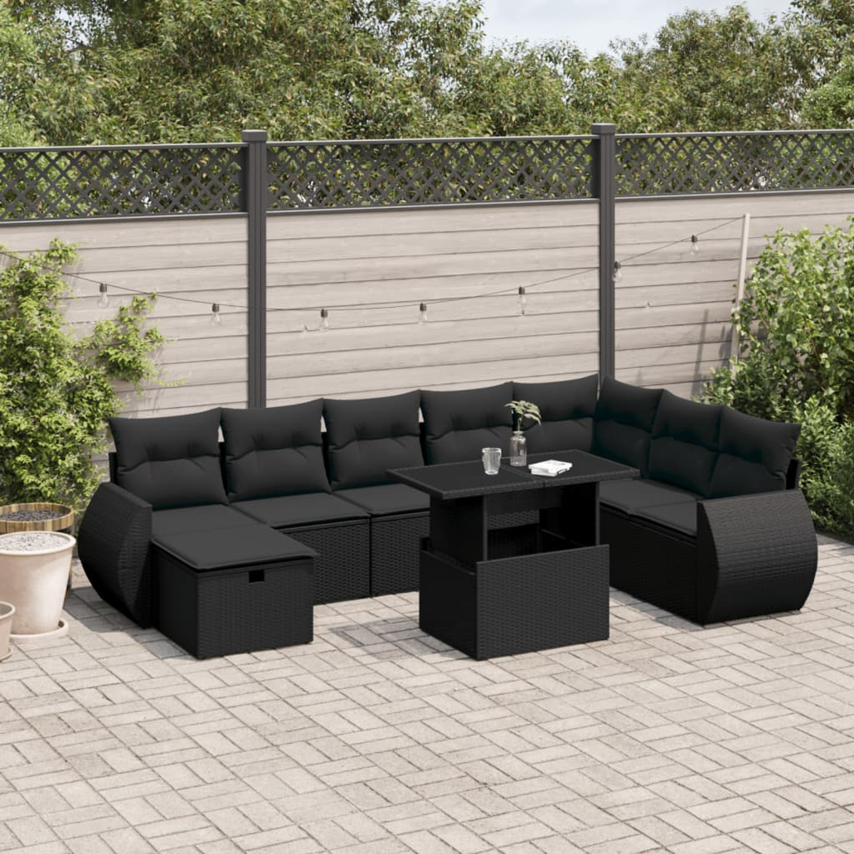 Schwarzes Ecksofa aus Korbgeflecht mit Tisch auf einer Backsteinterrasse. Hintergrund: Zaun und Grün.