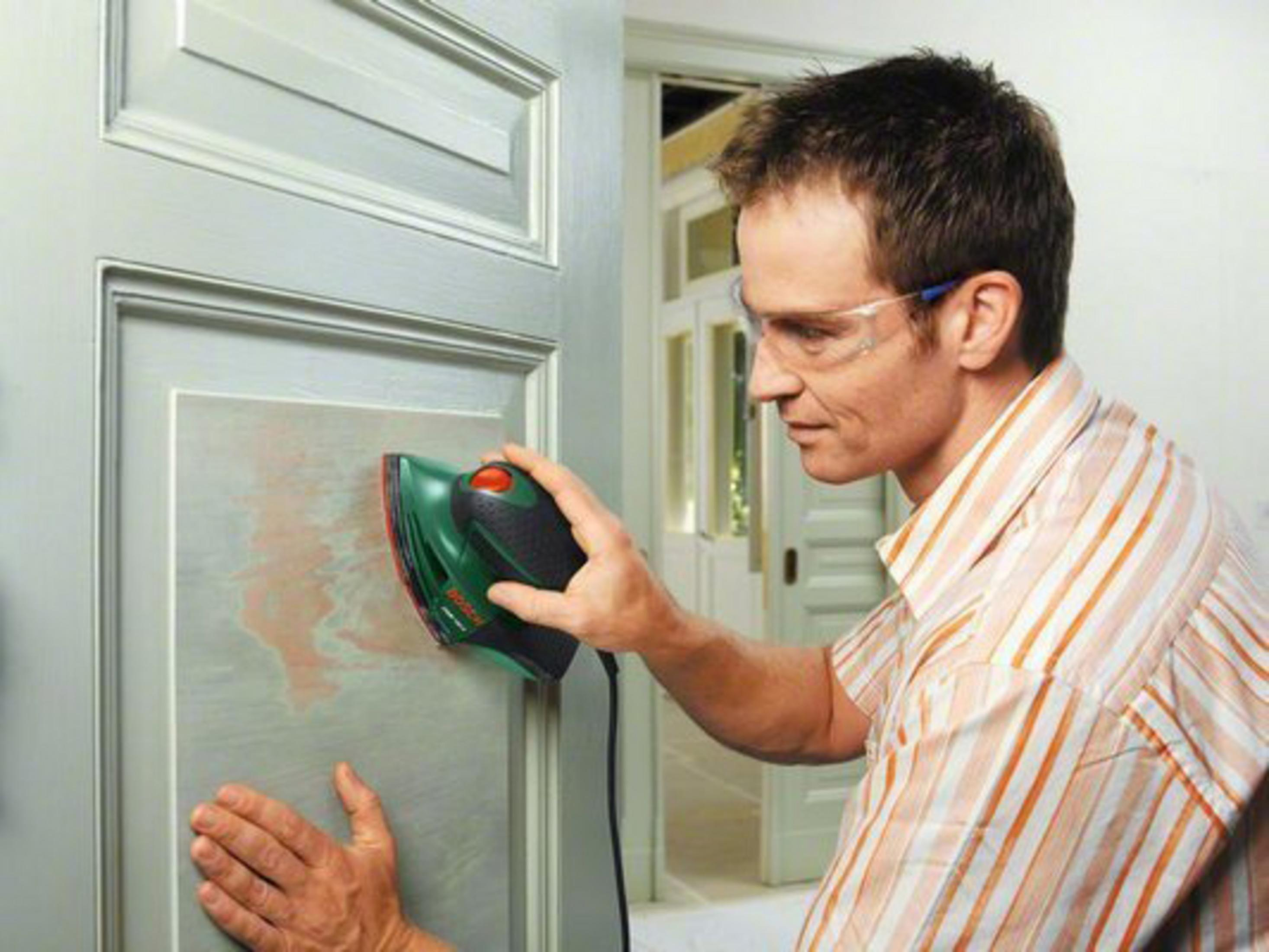 Homme utilisant une ponceuse verte sur une porte gris clair, portant des lunettes de sécurité.