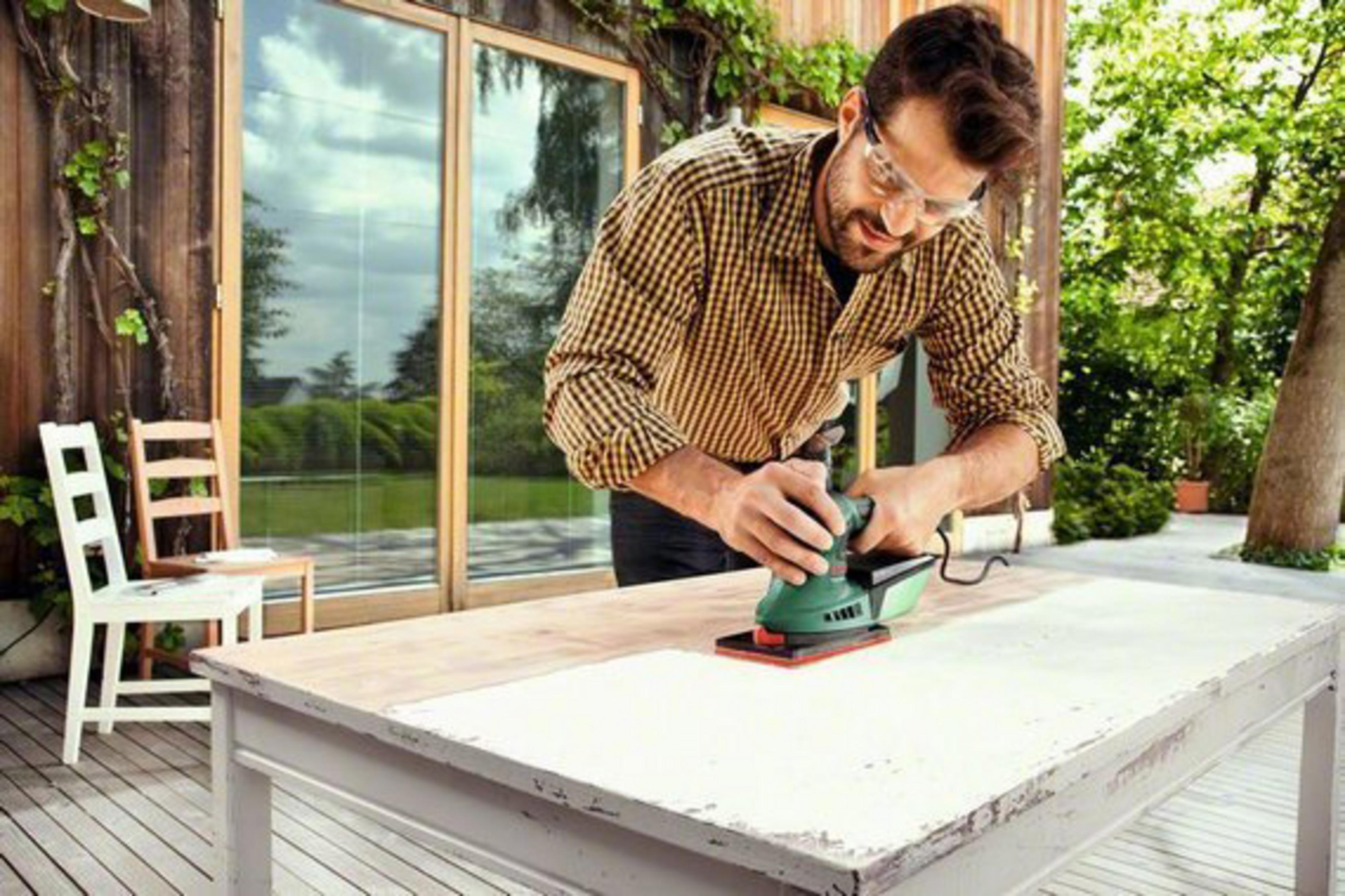 Un homme ponce une table en bois à l'extérieur. Une chaise blanche est à côté.