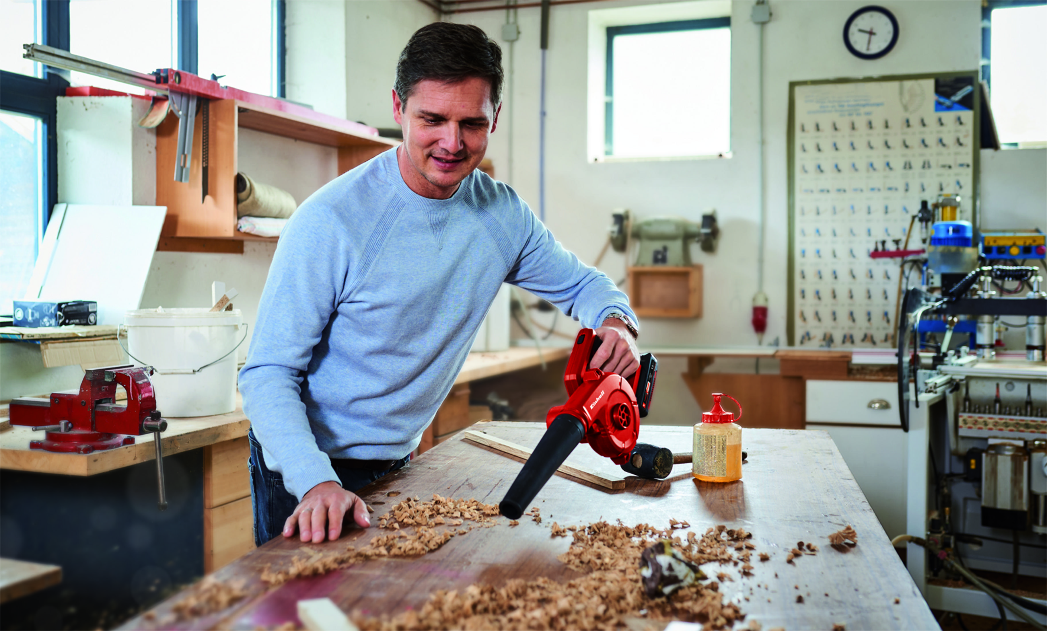 Mężczyzna używa czerwonej dmuchawy do liści na drewnianym stole pokrytym wiórami. Jest w warsztacie.
