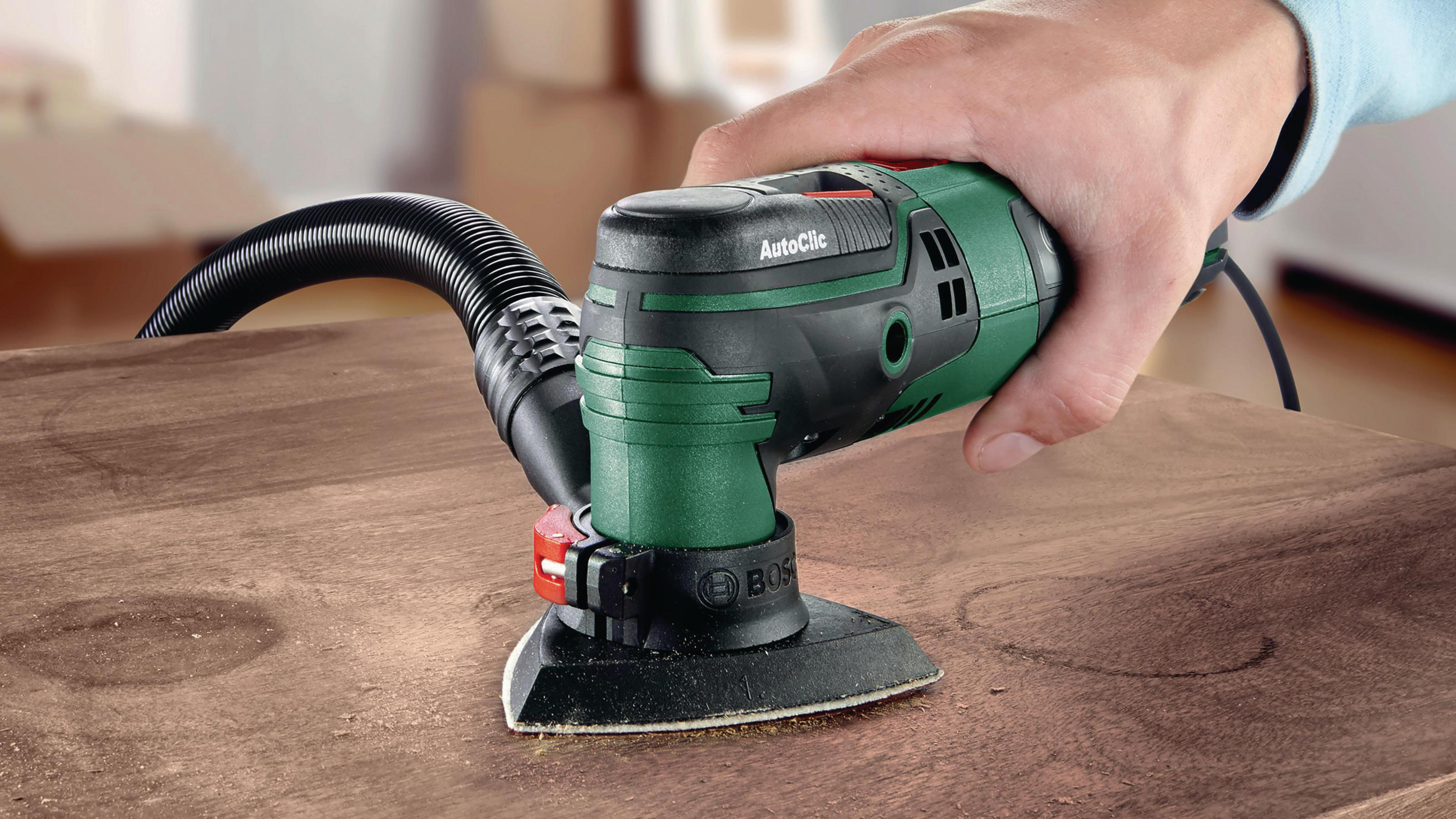 Mano che leviga una superficie di legno con una levigatrice verde e nera. L'utensile riporta la scritta 'AutoClic'.